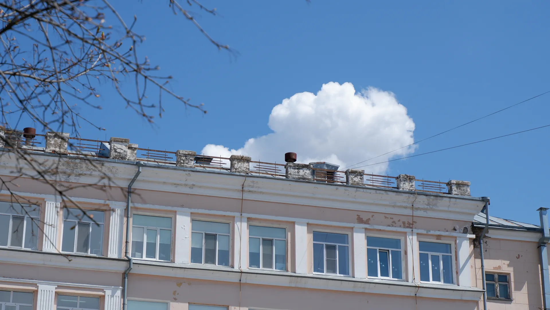 The top of a multi-story building showing some ventilation ducts and guardrails. The building's exterior walls are light-colored with multiple windows, under a sky with a few white clouds, partially obscured by tree branches.