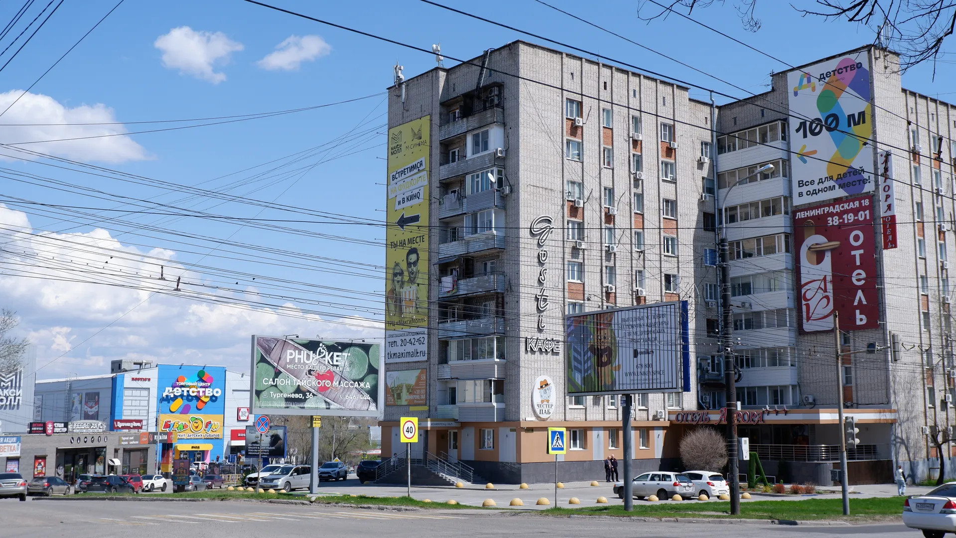 A street lined with multi-story buildings adorned with billboards and signs. Vehicles are driving on the road with traffic signs visible, under a sky with a few white clouds and crisscrossing power lines.