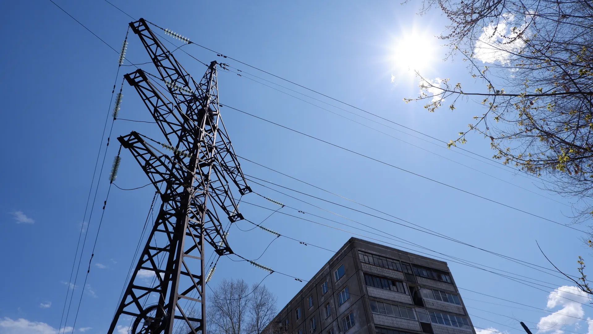 A complex metal electricity tower structure with multiple power lines extending from it. The sky is clear and the bright sun illuminates the tower and surrounding buildings, whose windows reflect the sunlight.