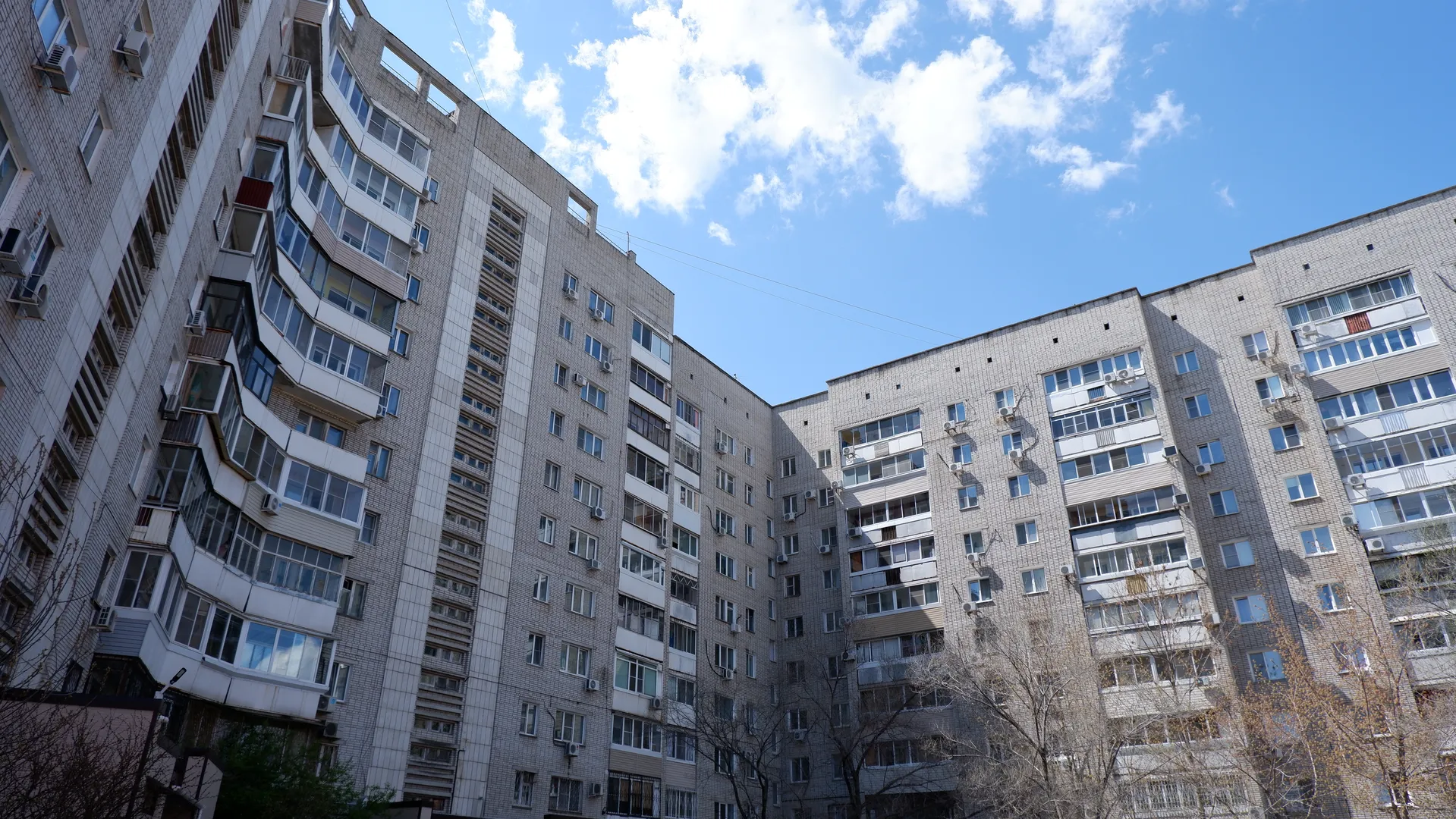 Two high-rise buildings forming a U-shaped structure, featuring grey tiled exterior walls and numerous windows. A few trees with sparse leaves stand between the buildings under a sky with several white clouds.