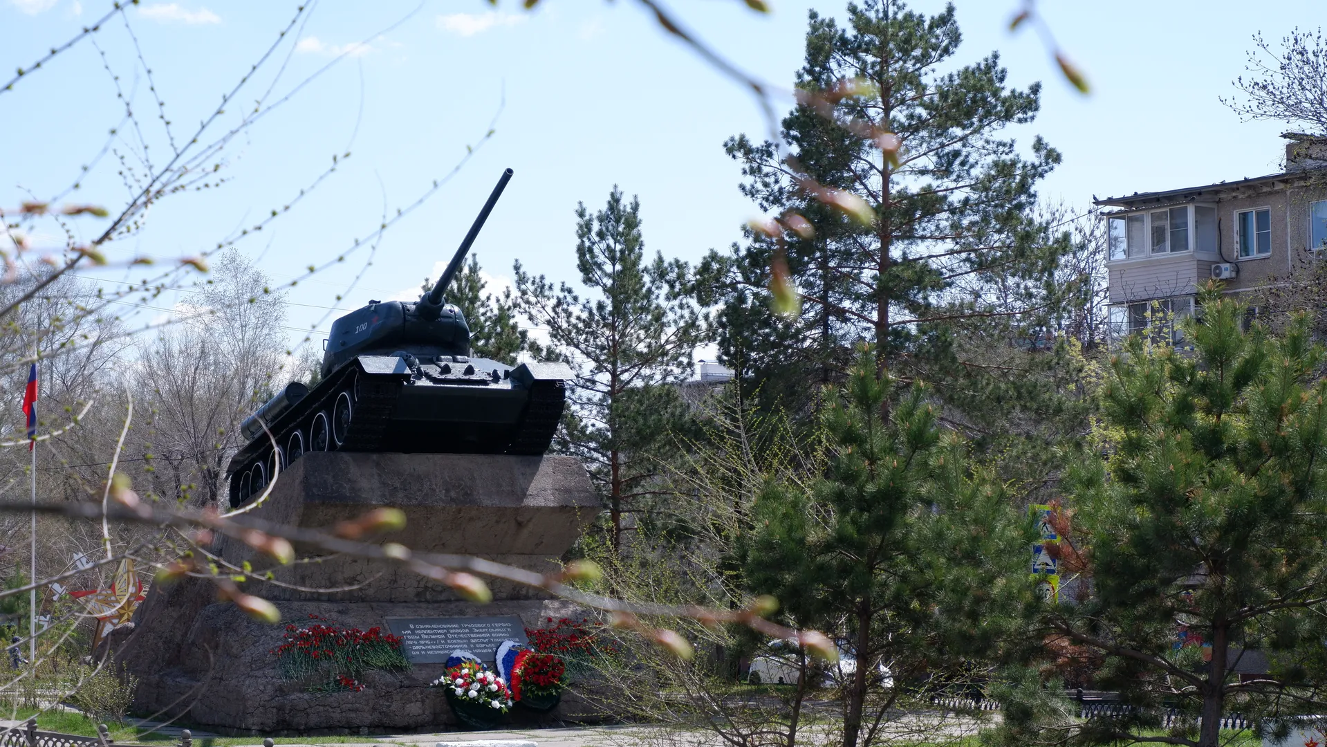 A tank monument stands atop a flower bed, with the tank's barrel pointing towards the sky. The monument is surrounded by several tall pine trees with dense foliage, against a background of a building with bright windows.