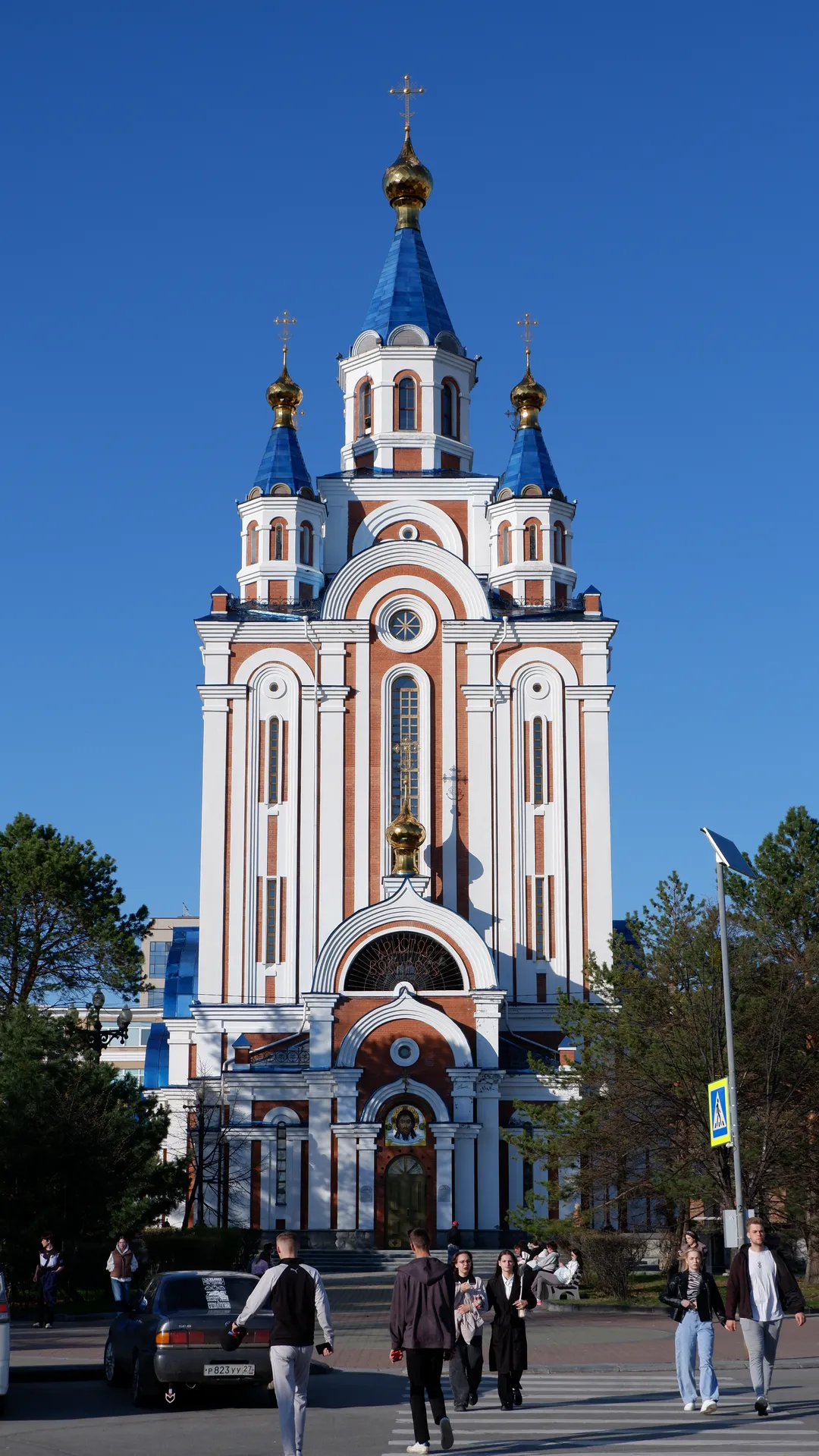 A church with golden domes and a blue roof stands under a blue sky. The front facade features multiple arched windows and decorative pillars, with people walking on the square ahead.