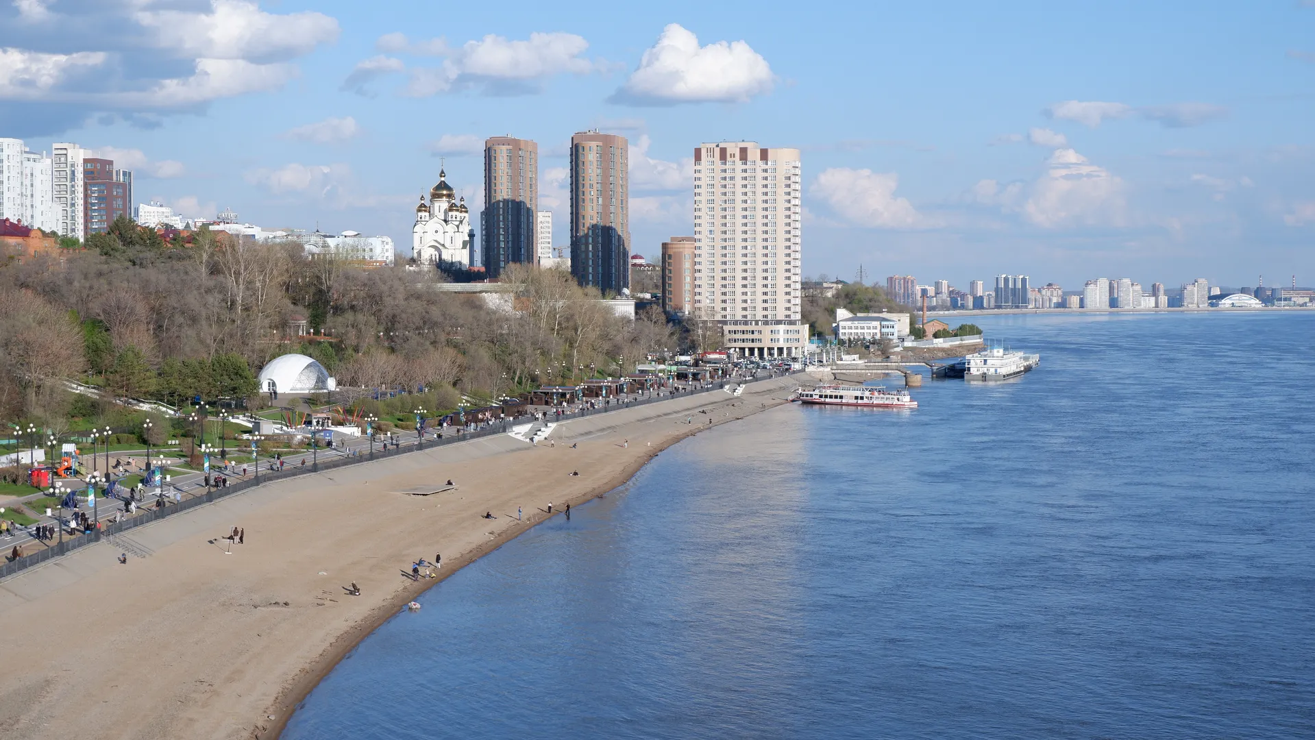 A sandy beach next to a river, featuring pedestrians and facilities. High-rise buildings and a golden-domed church can be seen in the distance under a sky with a few white clouds.
