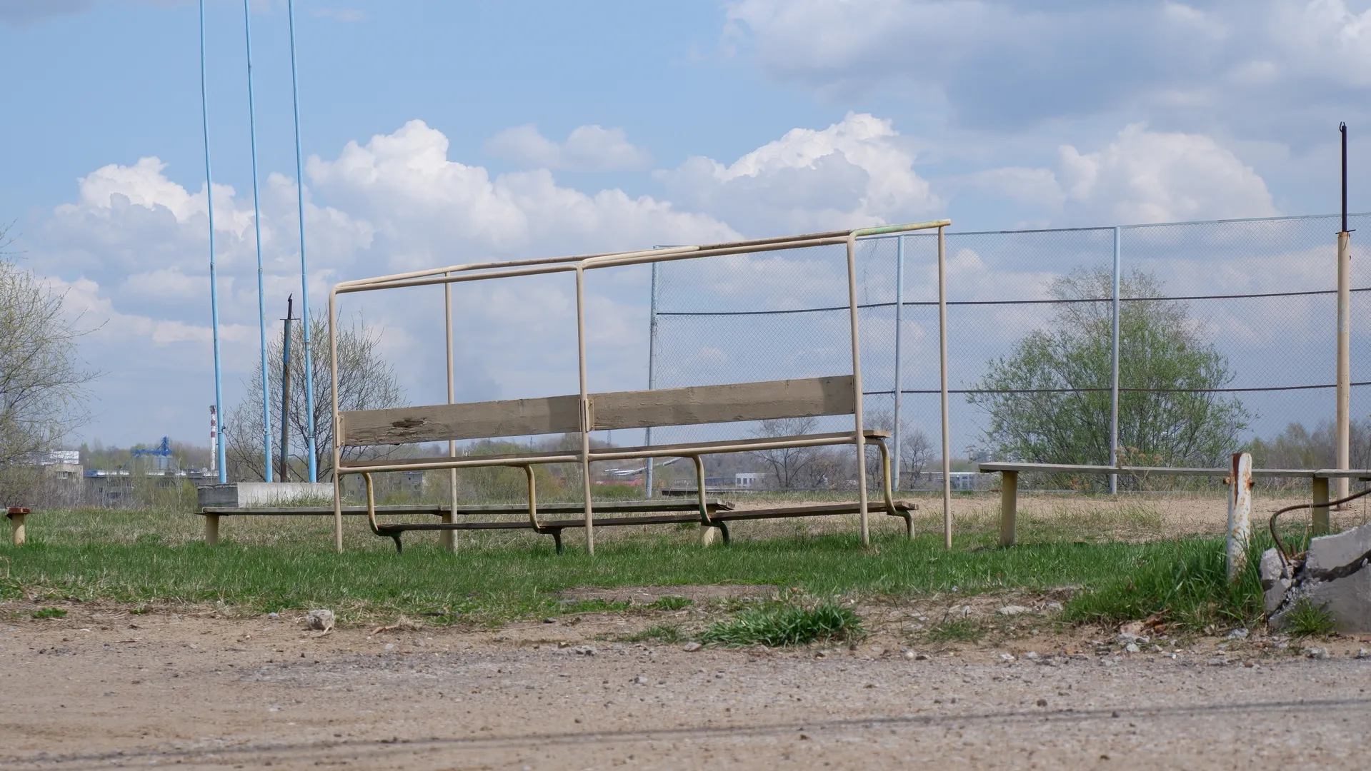 An open area with several worn-out benches in the foreground. Behind the benches is a lawn and a chain-link fence, with some utility poles and buildings visible in the distance.