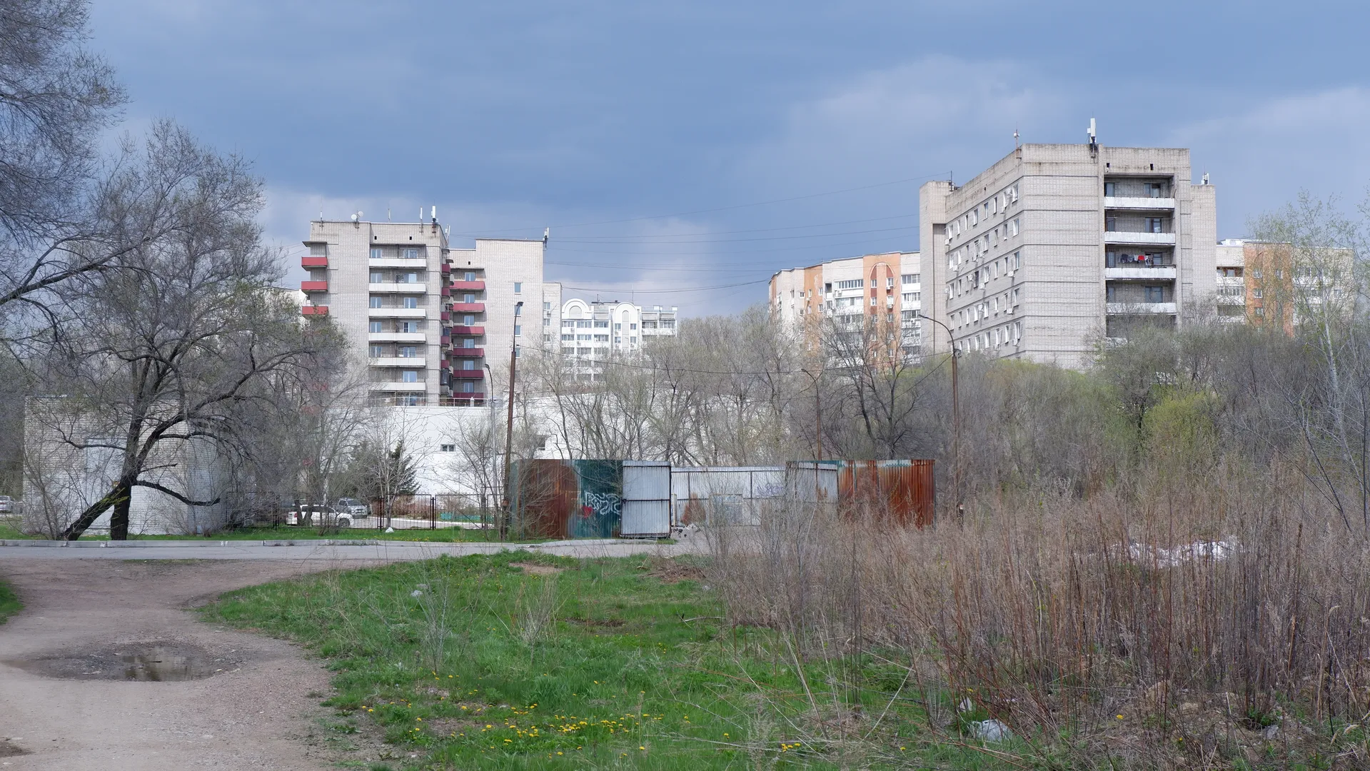 A multi-story building complex with trees and bushes behind it, fronted by a lawn. The buildings feature colorful exterior walls and neatly arranged windows under a cloudy yet well-lit sky.