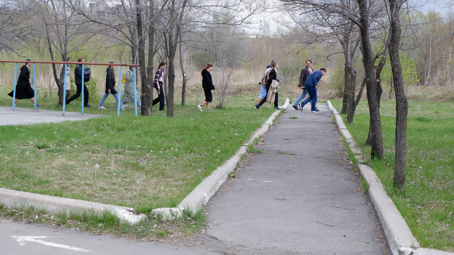 Several people walking along a park path flanked by trees and lawns. In the distance, some people can be seen standing next to a metal frame, against a background of buildings and open fields.