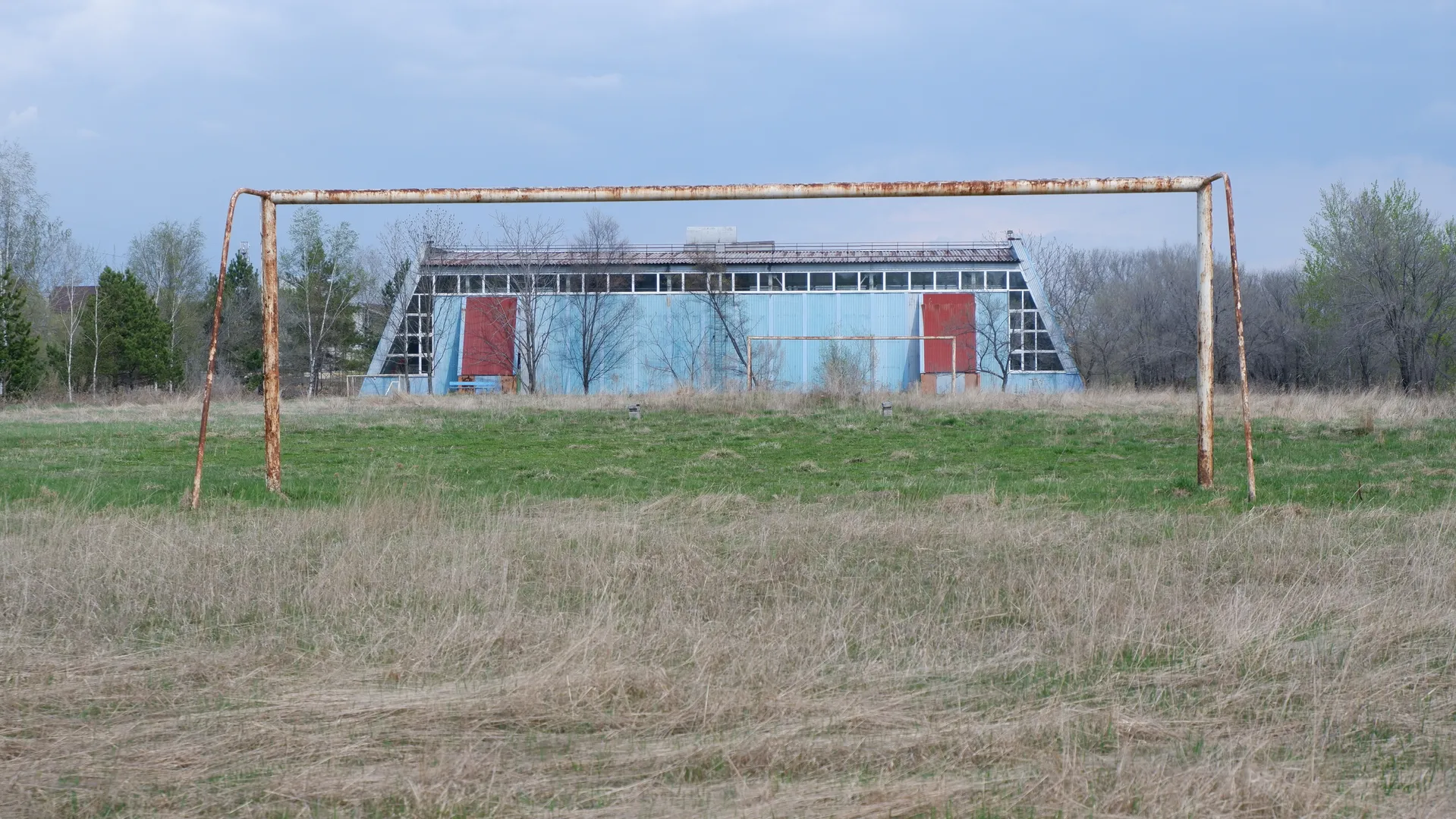 A rusty football goalpost stands on a patch of dried grass. In the background is a blue building with two red doors, with trees and the sky visible in the distance.