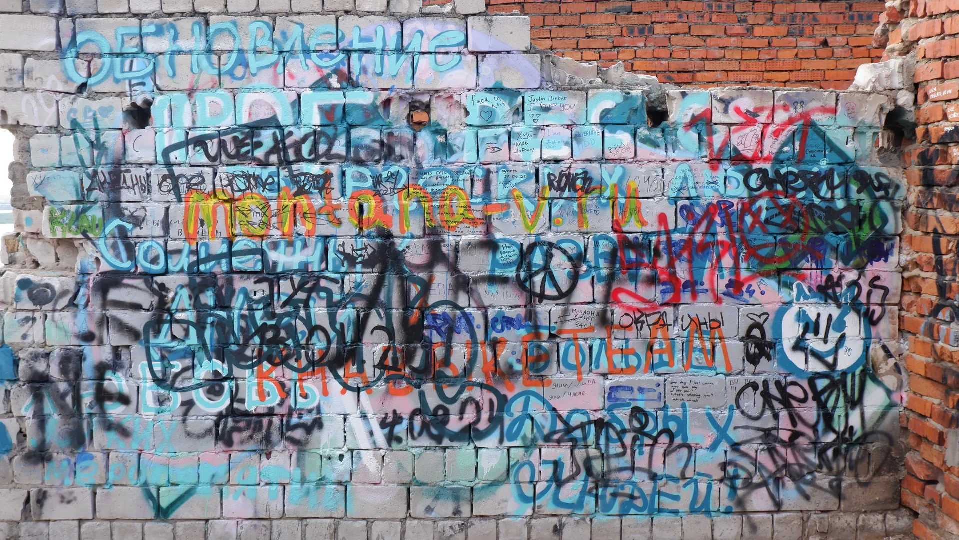A white brick wall covered in colorful and intricate graffiti, featuring numerous letters, symbols, and simple shapes. The wall is partially damaged, exposing the texture of the bricks.