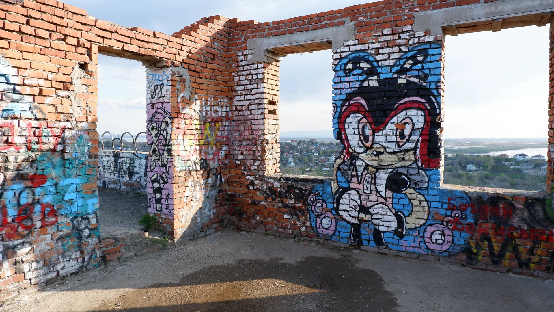 Large cartoon graffiti depicting a character wearing a red headscarf is painted on the ruins of an abandoned building. The red brick walls and concrete structure are clearly visible, with a distant cityscape and a river seen through the gap.