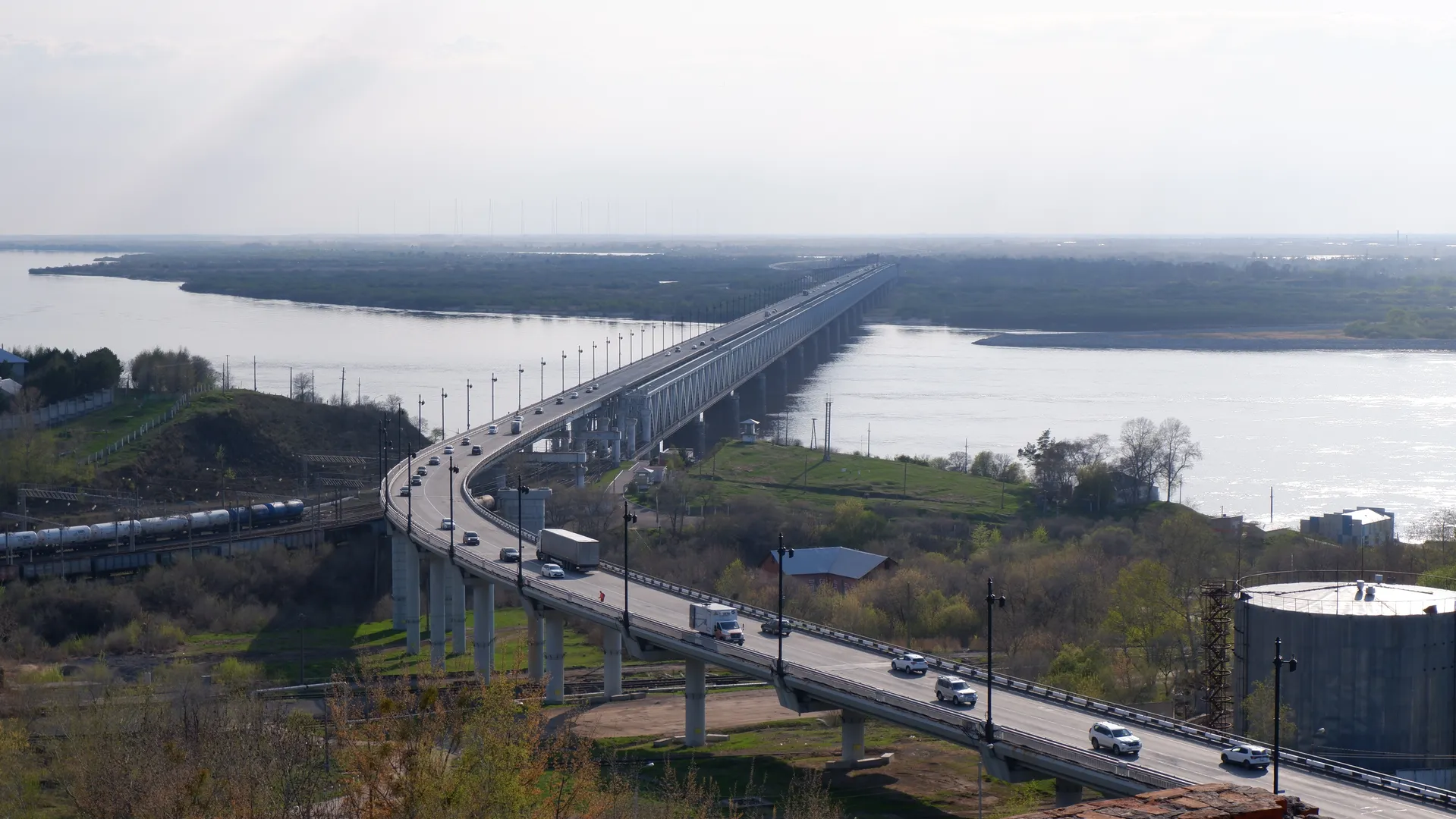 A long bridge with multiple lanes spans across a wide river, with vehicles driving on it. The bridge is equipped with streetlights and guardrails on both sides, with flat terrain and some vegetation visible in the distance.