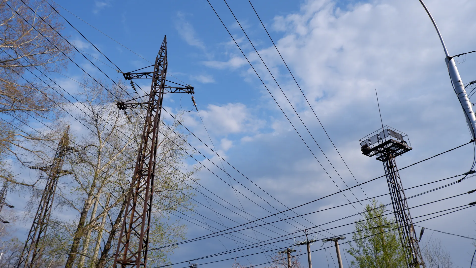 High-voltage power towers and multiple power lines stretch across a blue sky with white clouds, flanked by trees. The tower structure is complex and the wires are neatly arranged, with sparse clouds in the distant sky.