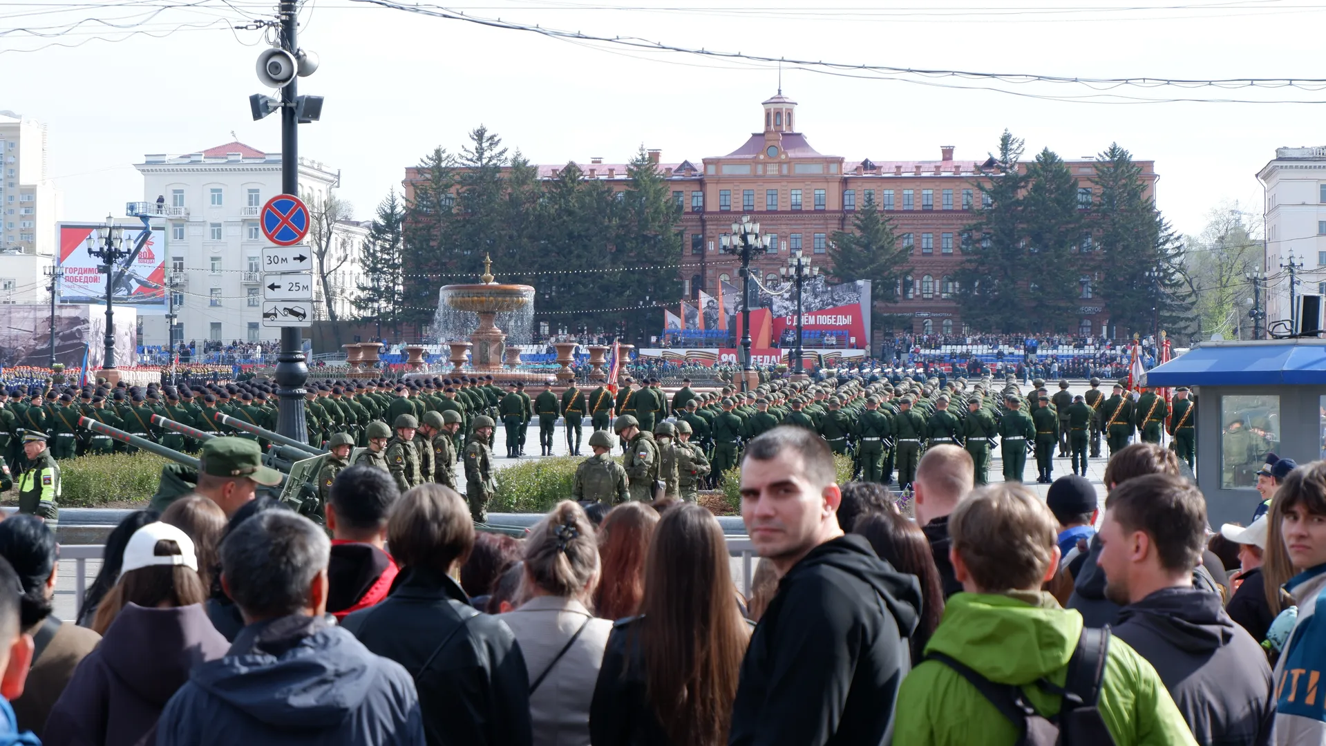 A large number of military personnel lined up on a square, with a fountain and spectators in front, against a background of red buildings and green trees. Some spectators wear black coats, while others wear white hats.