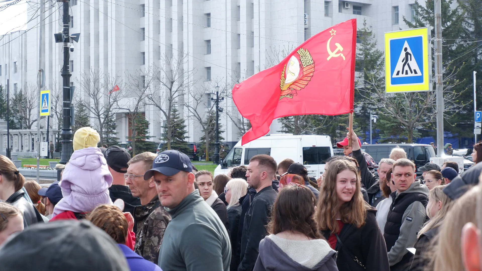 A person in the crowd holds a red flag aloft, featuring a hammer and sickle design, against a background of tall buildings and bare trees, with pedestrians wearing various coats.
