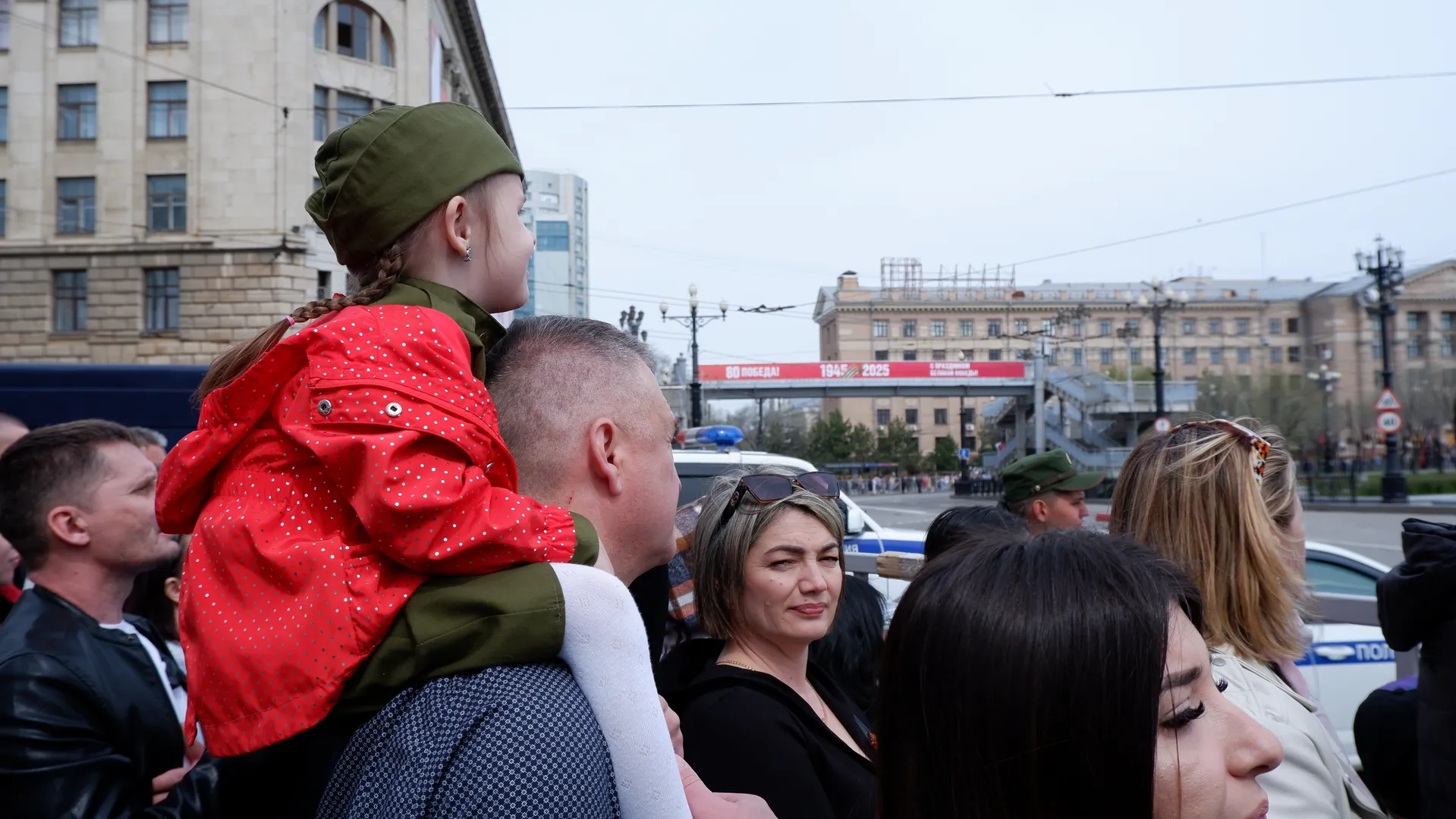 A child in a red coat sits on an adult's shoulders amidst a dense crowd. High-rise buildings and vehicles are in the background, and a woman wearing sunglasses smiles.