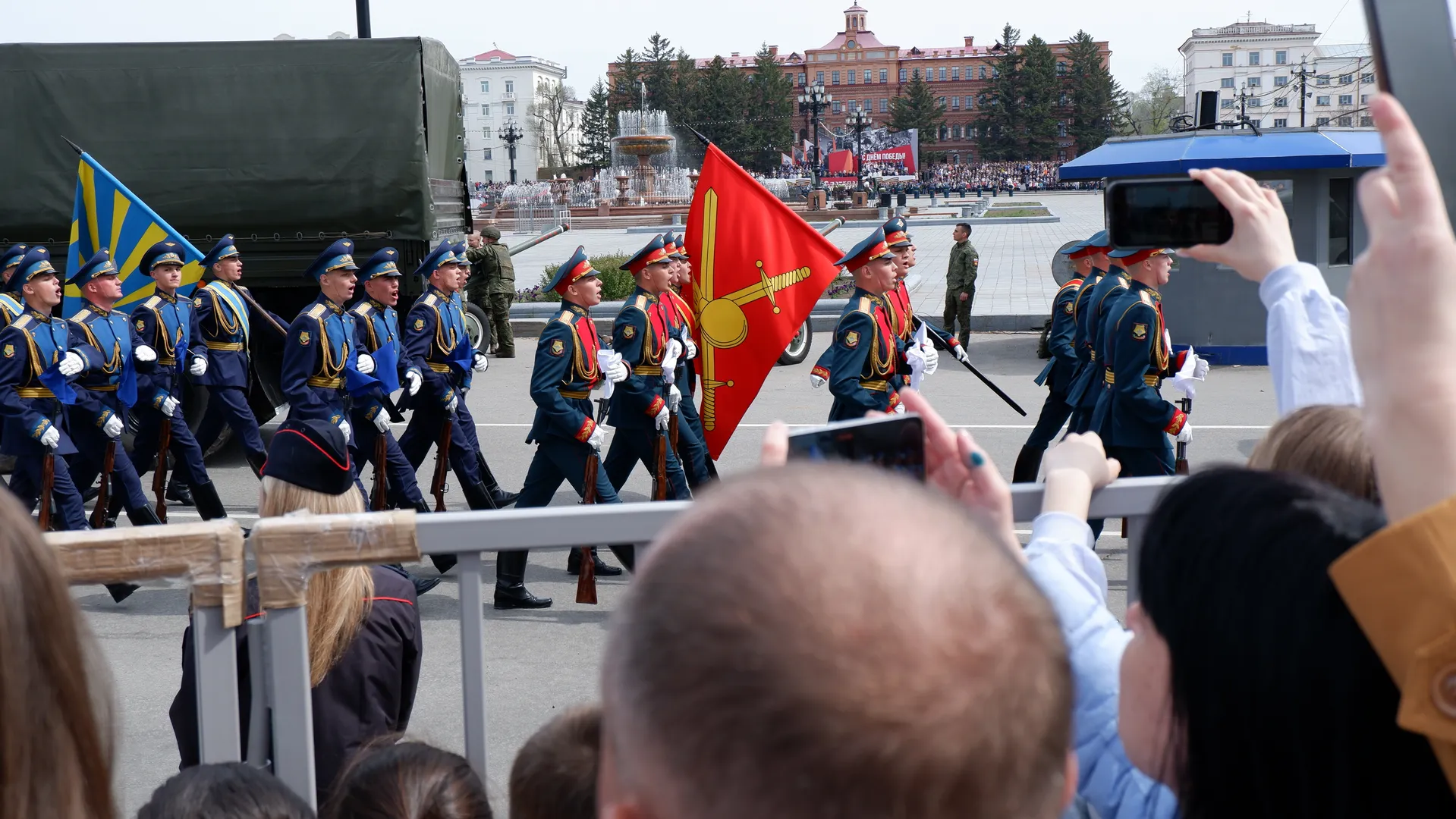 A group of military personnel in blue uniforms marching while holding flags and rifles, with a military truck and a fountain in the background. Spectators behind the guardrails film the scene with their phones.