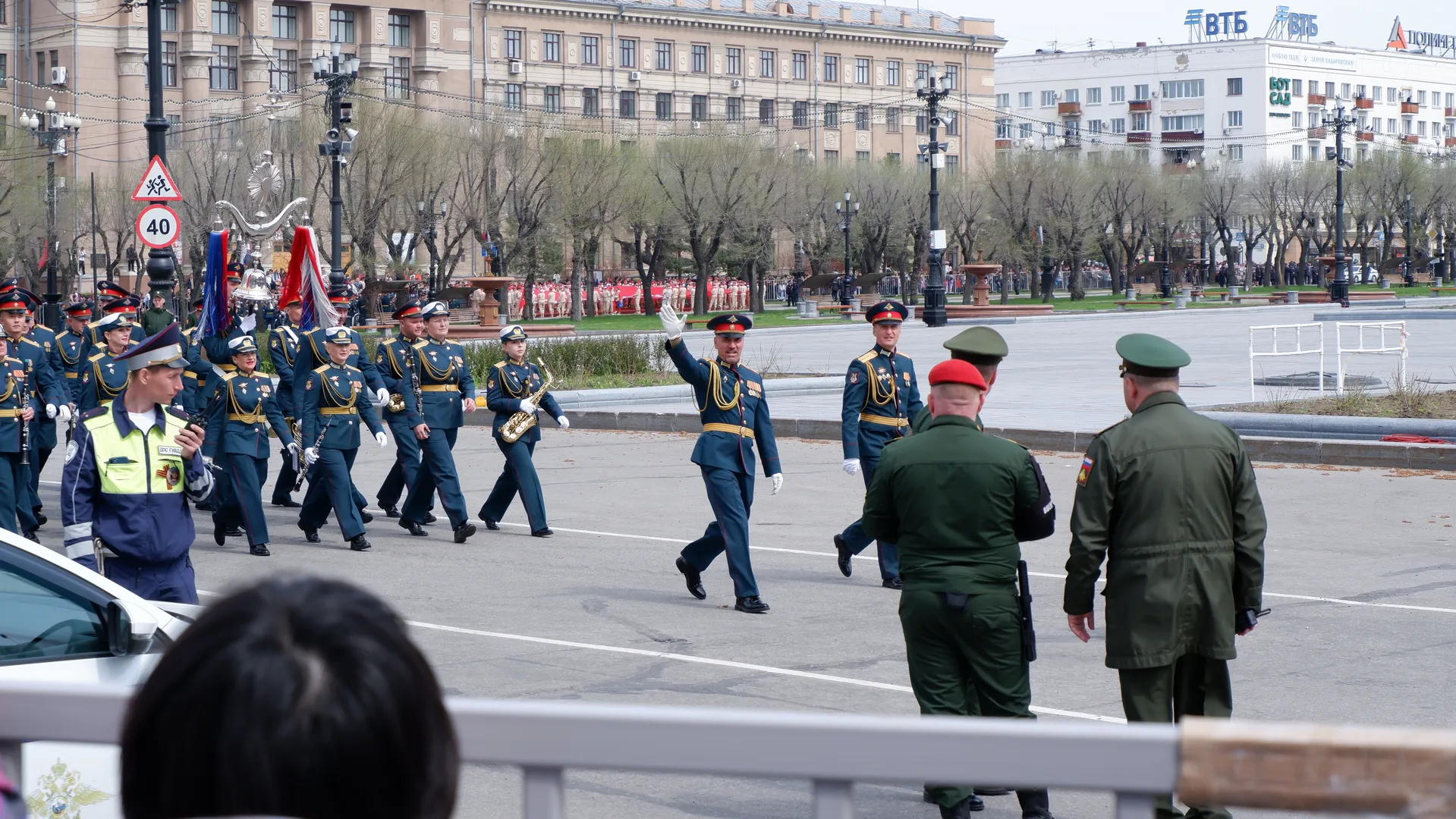 A military band marching, with members wearing dark uniforms and caps, fronted by two personnel in green military uniforms, against a background of city buildings and trees.