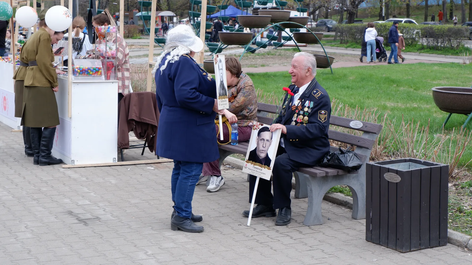 An elderly man in military uniform sits on a bench, with a woman in a blue coat standing beside him. A display board with photos and text stands in front of the elderly man, with amusement rides and pedestrians in the background.