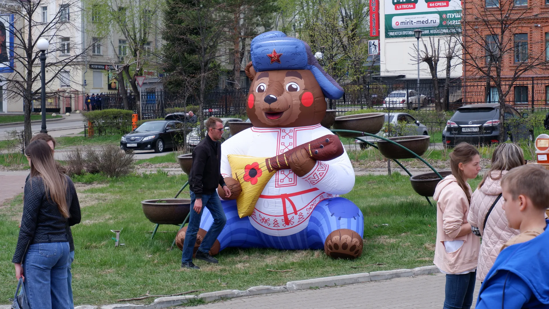 A massive inflatable bear sculpture situated on a lawn, wearing a blue hat with a red star and white clothing, with several people standing around watching against a background of trees, cars, and buildings.