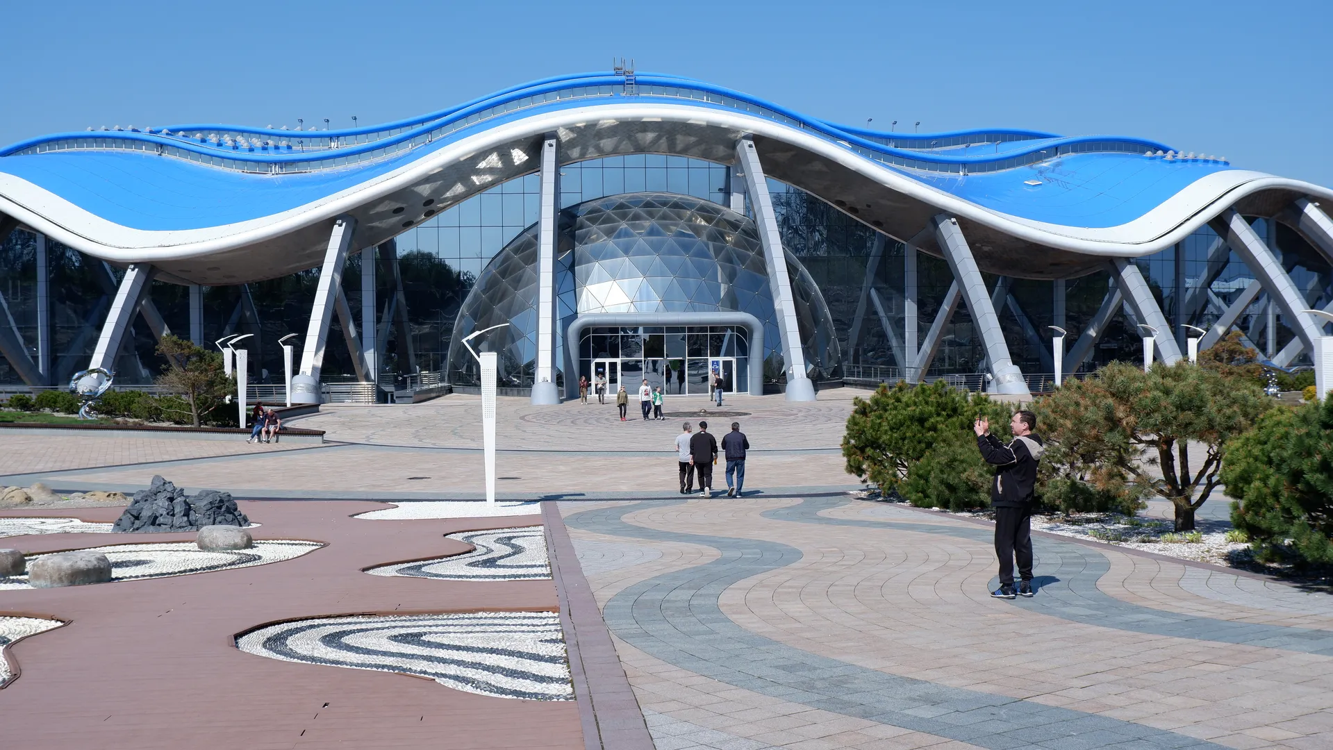 A modern building featuring a wavy blue roof and a glass curtain wall, with a few people walking on the square in front. The building entrance has a large spherical structure, surrounded by some planted trees, and the square ground is designed with curved patterns and stone decorations.