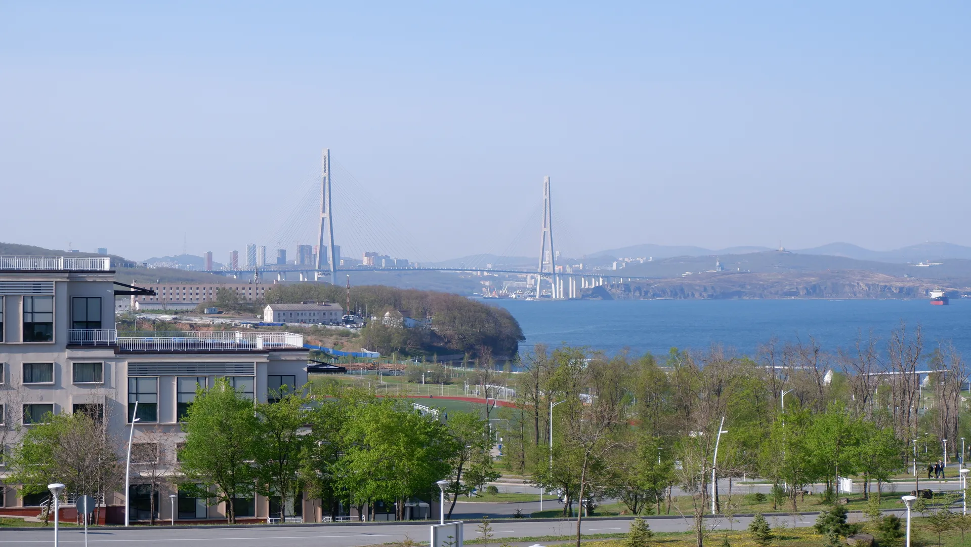 A cross-sea bridge visible in the distance, connecting the cityscapes on both shores, with trees and some buildings in the foreground over calm water under a clear, open sky.