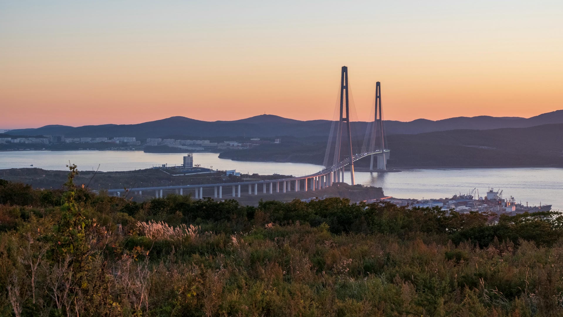 A bridge spanning across a bay at sunset, with the sky showing an orange-yellow gradient. The bridge connects both shores, with rolling mountains in the distance.