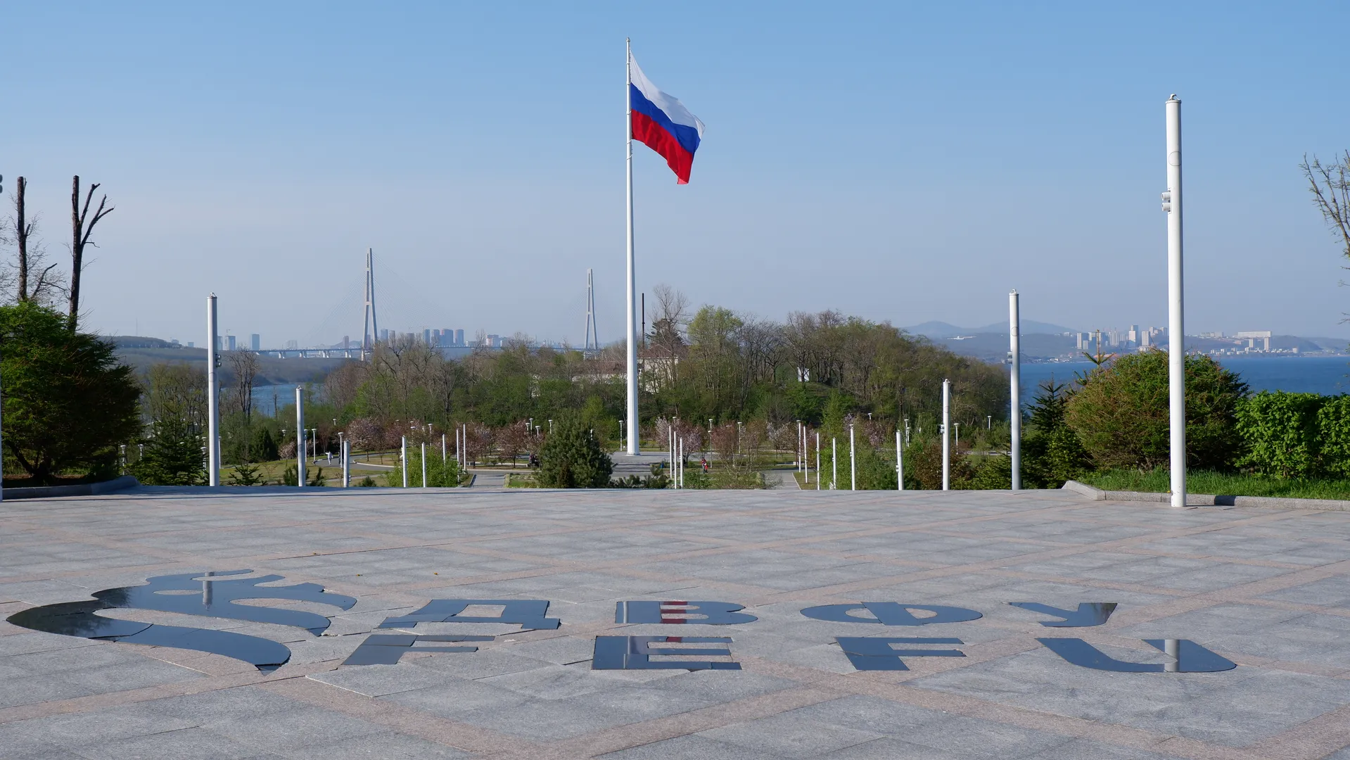 A square paved with large stone bricks, with a tall flagpole standing in the center flying the Russian national flag. The capital letters 'ДВФУ' are inscribed on the square ground, surrounded by green plants and trees, with the cross-sea bridge and city skyline visible in the distance.