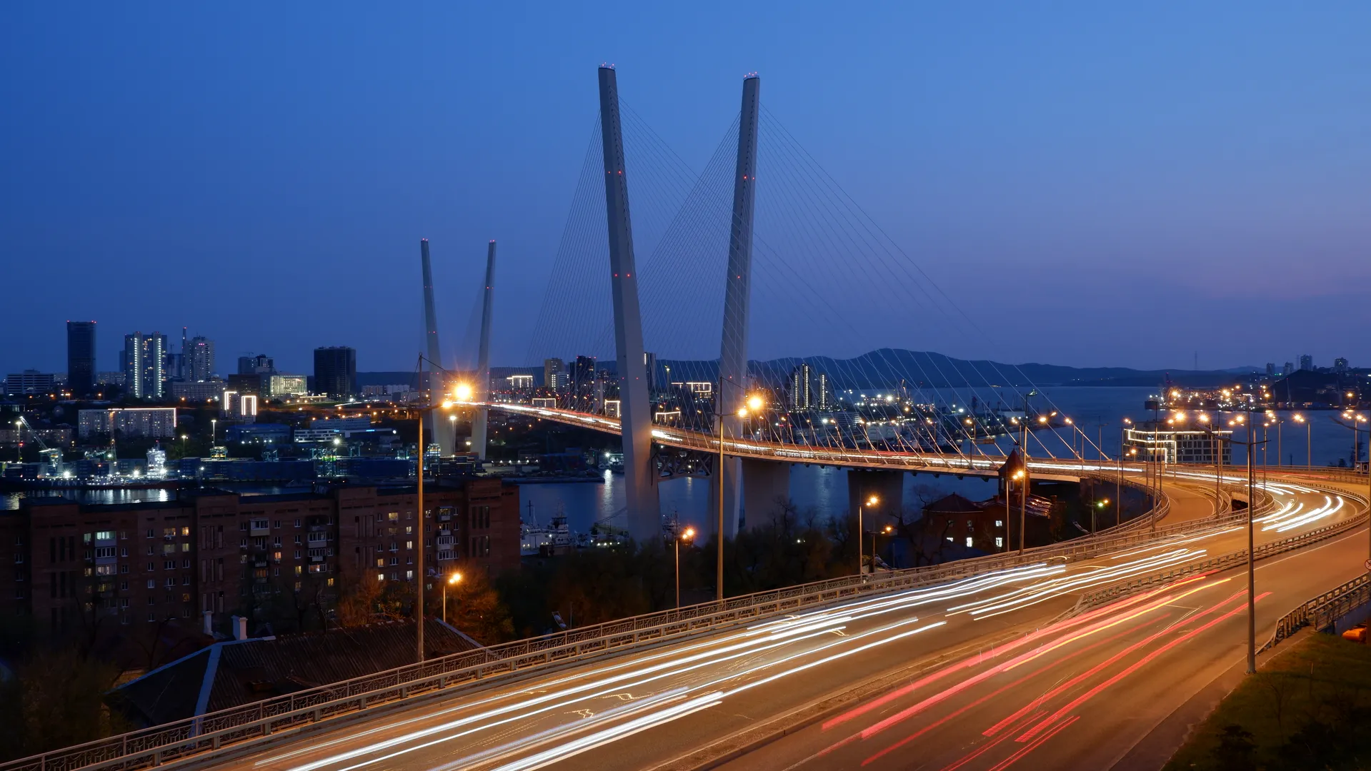 A city bridge brilliantly illuminated at night, with light trails left by moving vehicles. The bridge features a cable-stayed design with towering pylons, against a background of distant city lights and mountain contours.