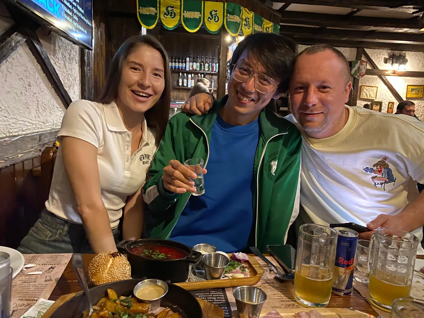 Three people sitting at a dining table; the person in the middle wears a green coat over a blue inner top, holding a glass of transparent drink. The person on the left wears a white short-sleeved shirt, and the person on the right wears a patterned white T-shirt. Food, drinks, and tableware are placed on the table, with bottles and decorative flags in the background.