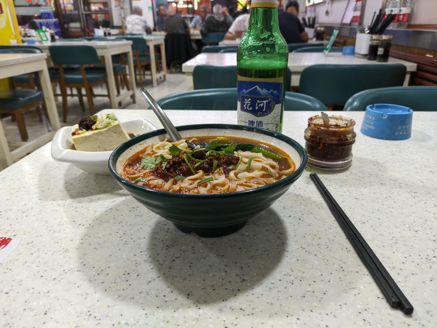 A bowl of noodles, a bottle of beer, a dish of tofu, and chili sauce on the table.
