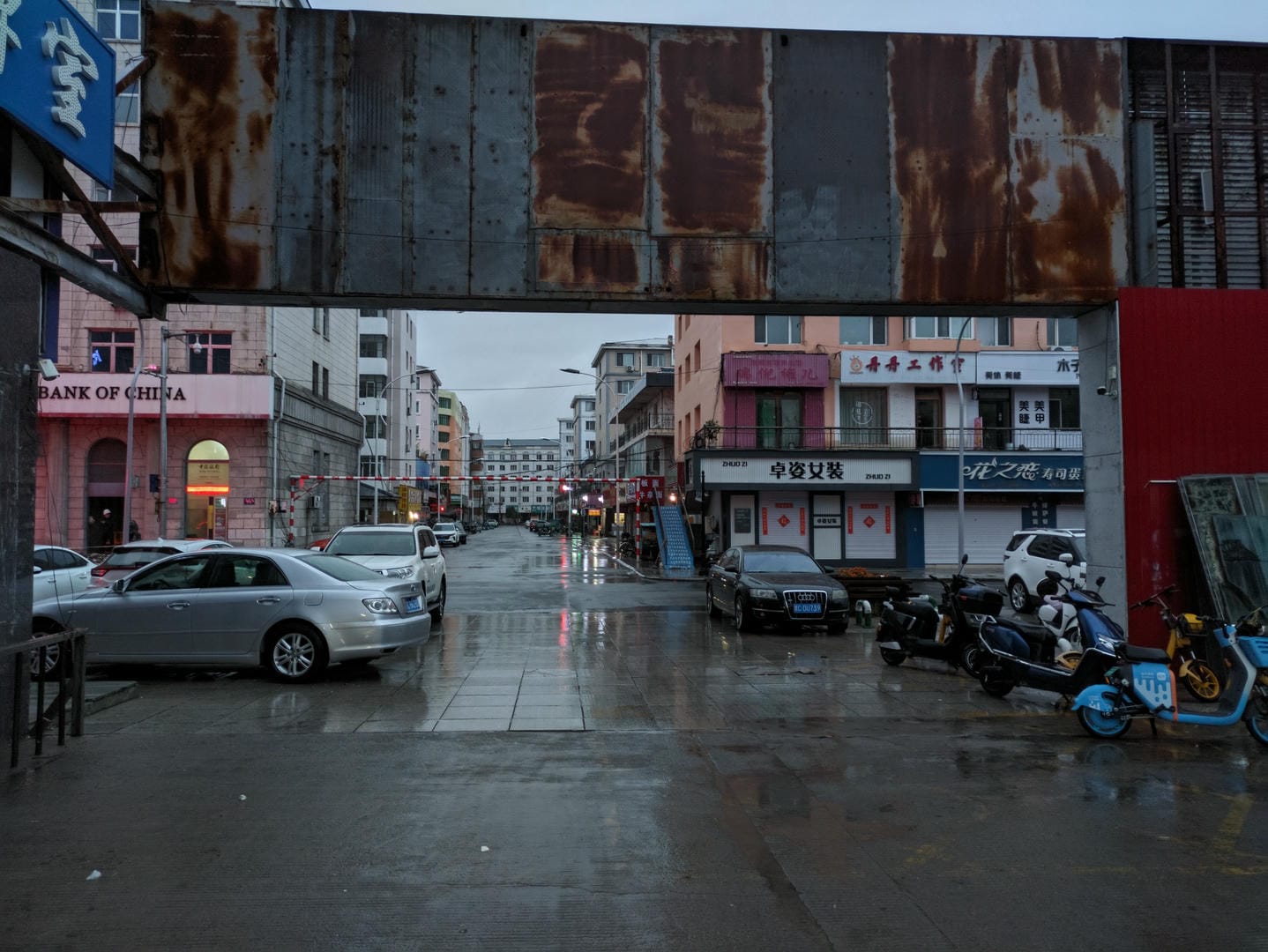 A wet and slippery street after the rain, with cars and motorcycles parked on both sides, and a rusty metal structure overhead.