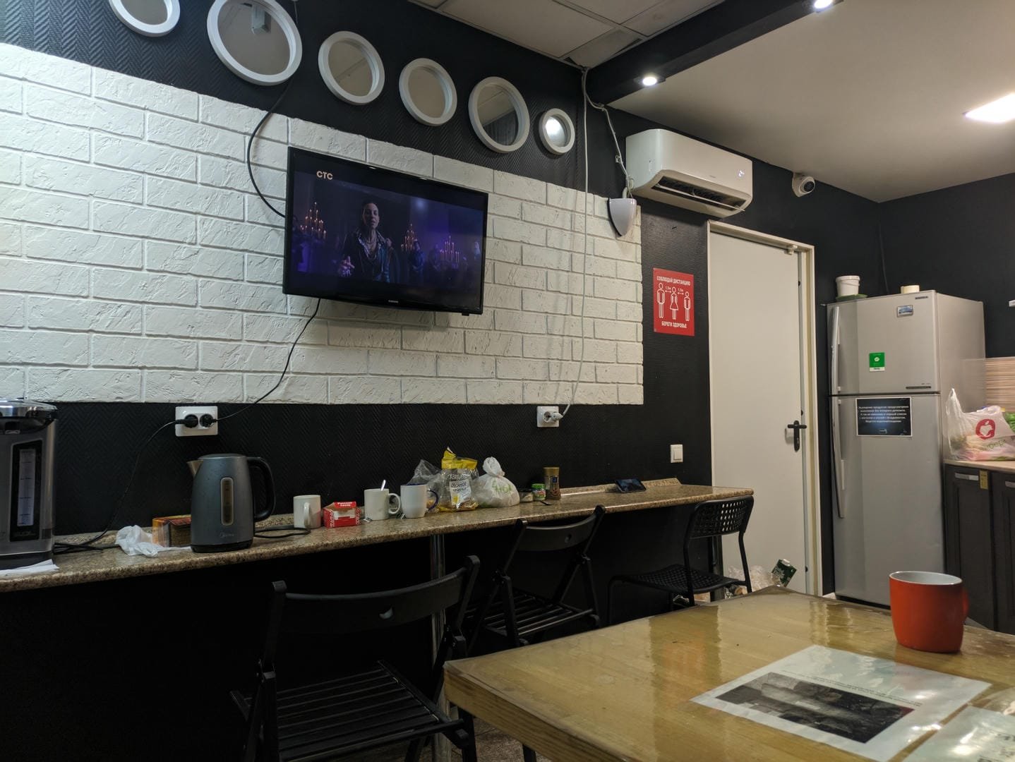A kitchen area with a TV playing on the wall, a kettle, cups, and other items on the countertop, and a refrigerator on the right.
