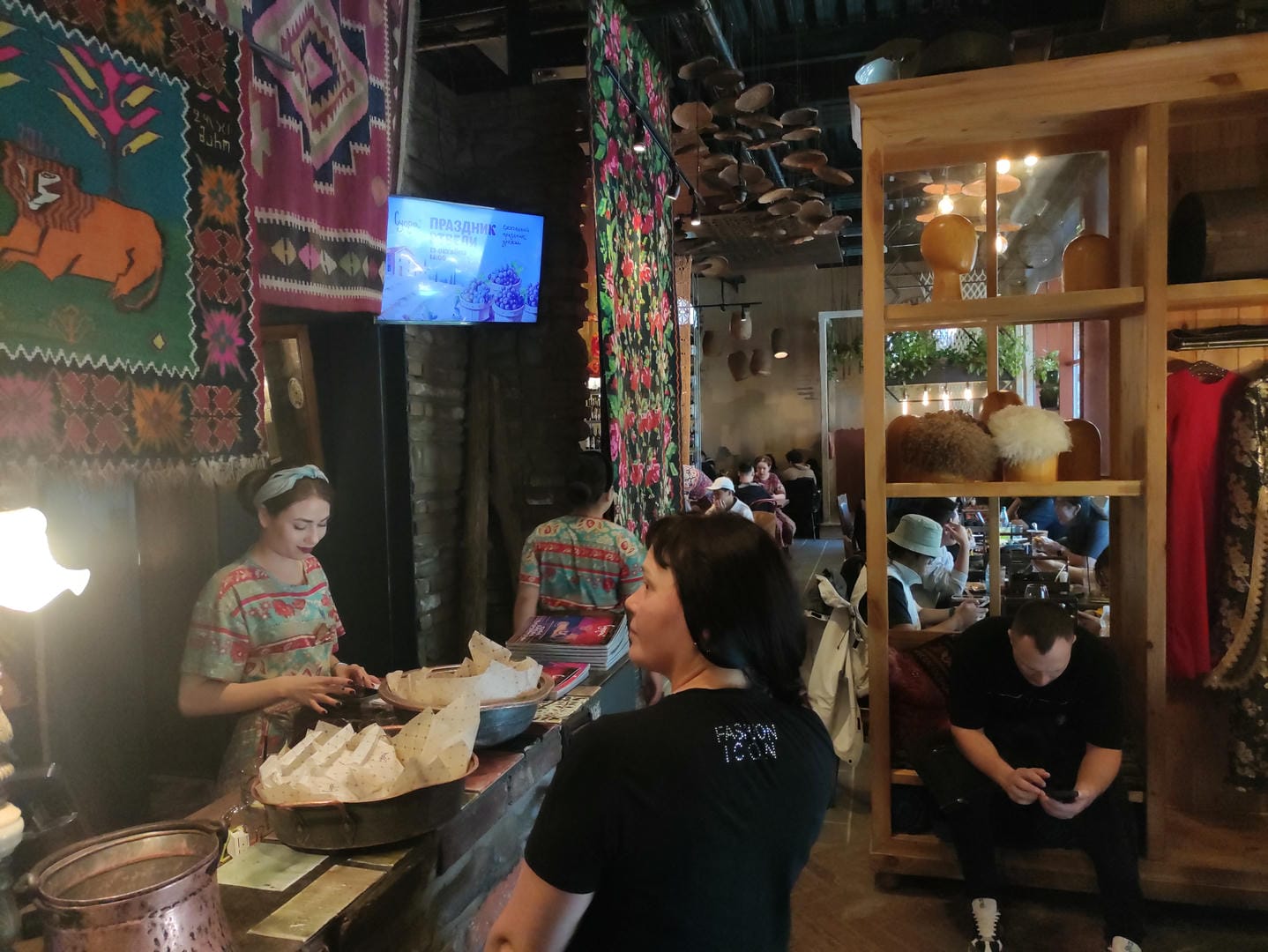 The interior of a restaurant decorated with colorful tapestries, where a waiter prepares food and customers sit at tables.