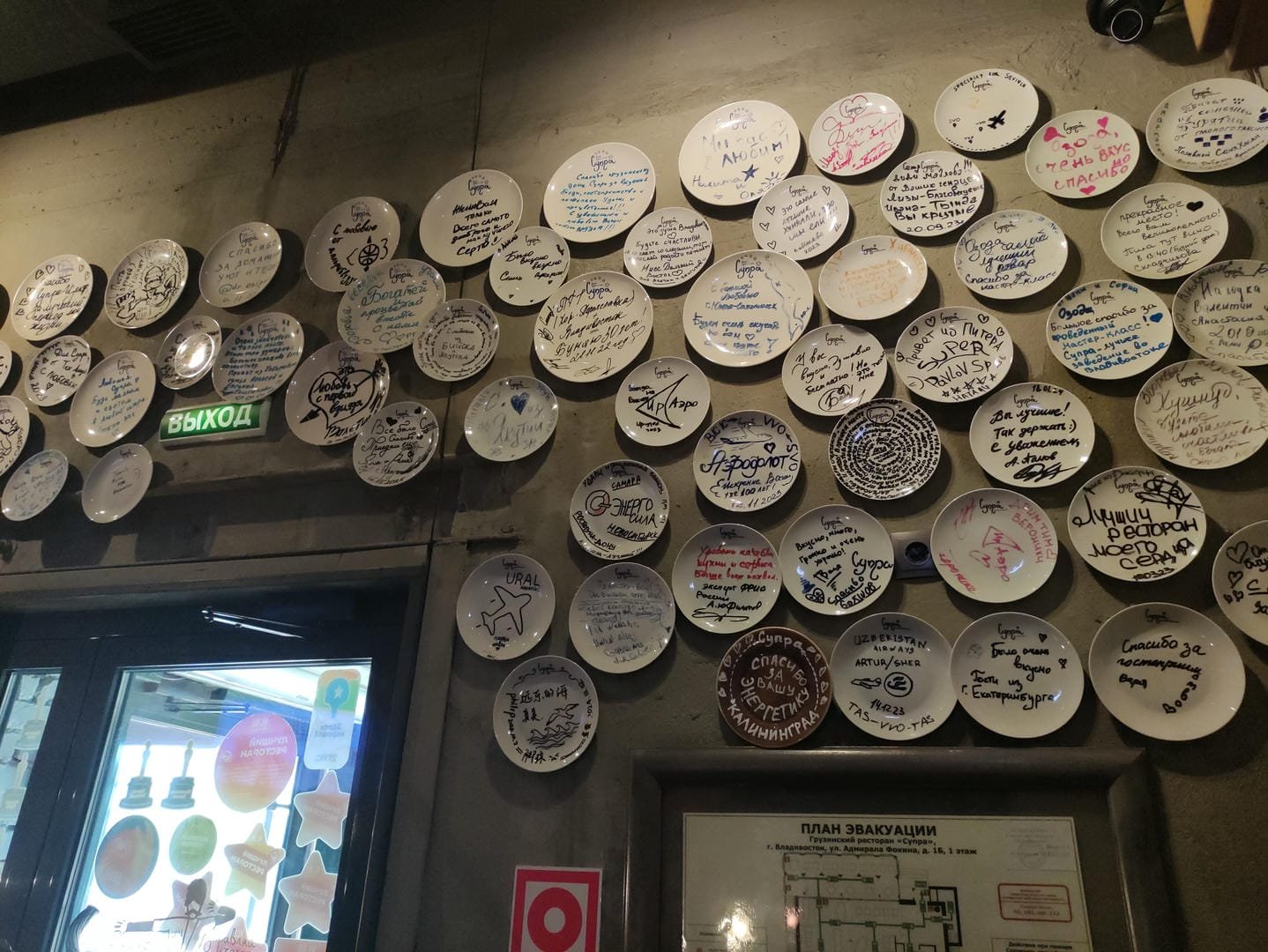 A wall covered with white plates featuring handwritten messages, with a green sign above the doorway.