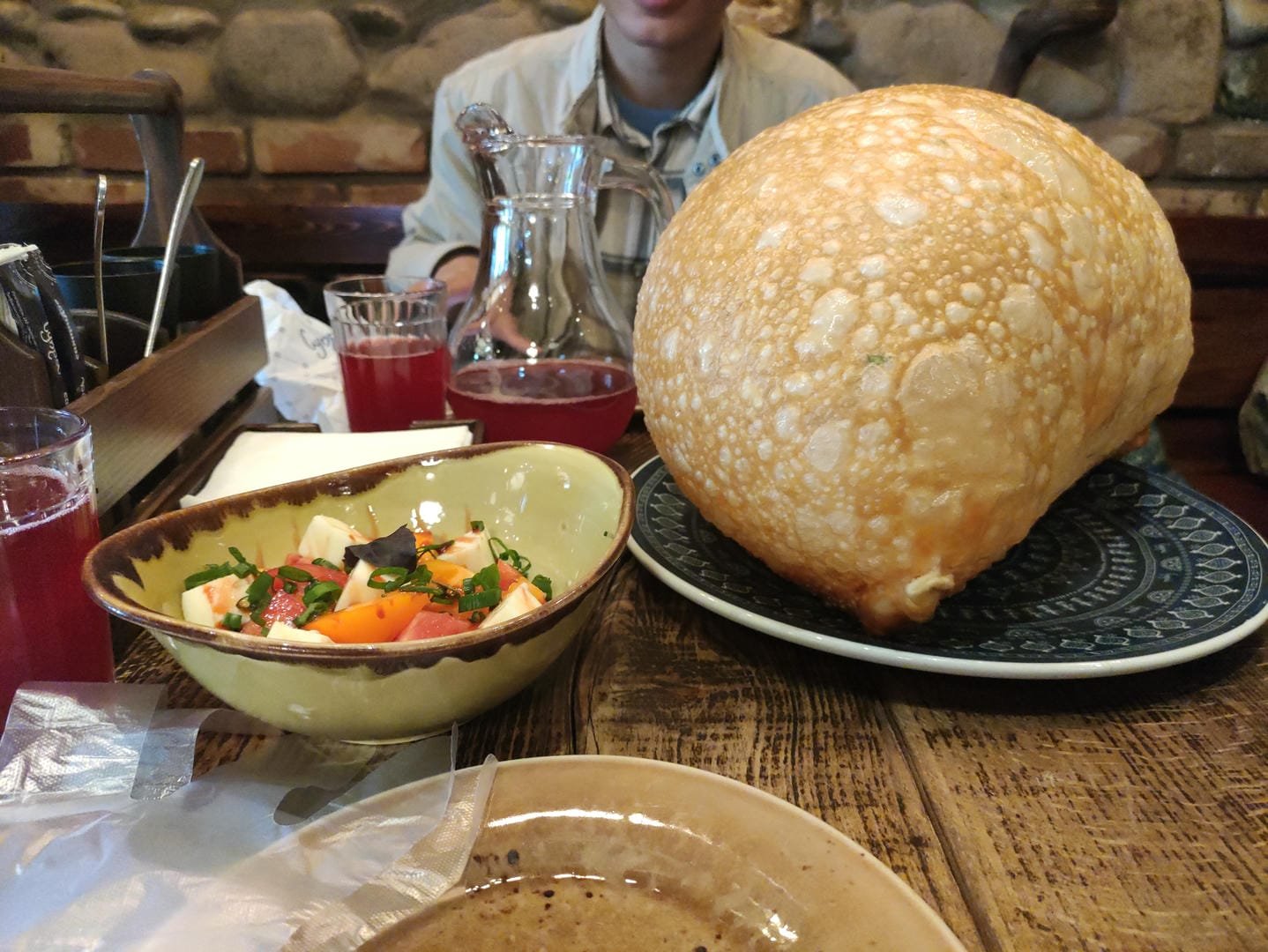 A large loaf of bread, a bowl of salad, and a glass of red beverage on a dining table, with people seated in the background.