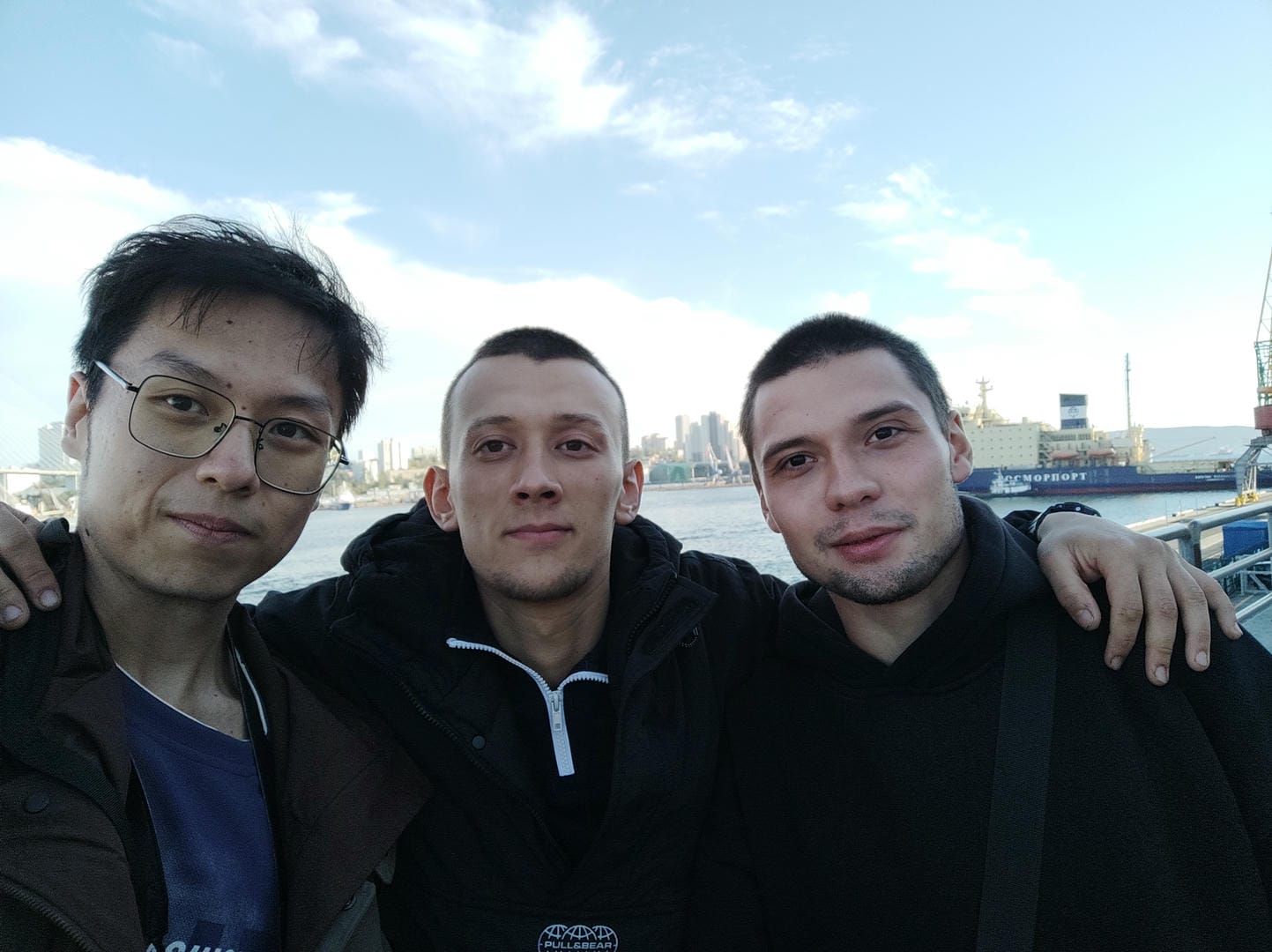 Three people posing for a photo outdoors, with the water surface and distant city buildings in the background.