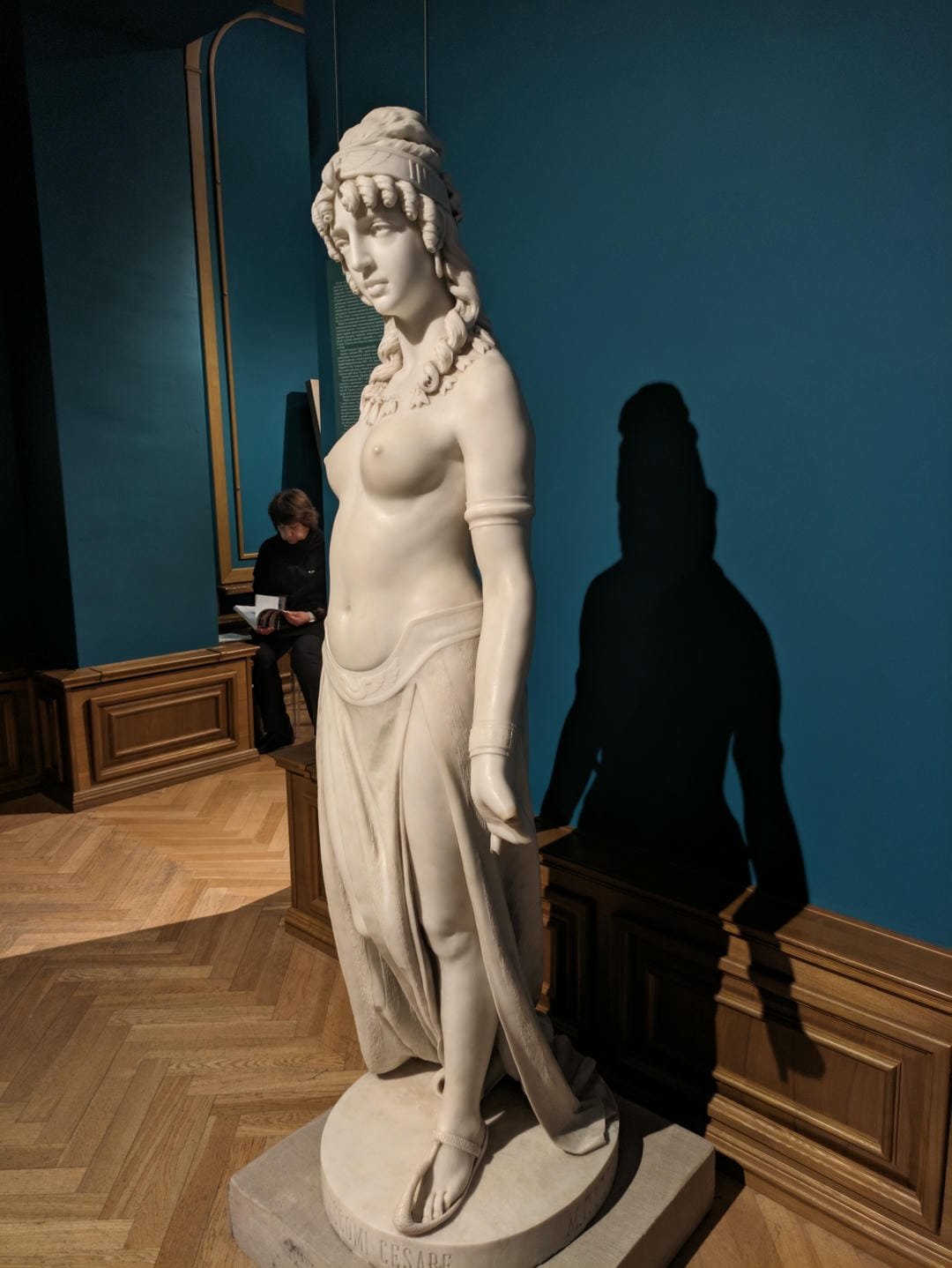 A white marble statue of a semi-nude woman wearing a headscarf, with her right hand on her chest, her left hand hanging down naturally, and a pair of sandals at her feet.