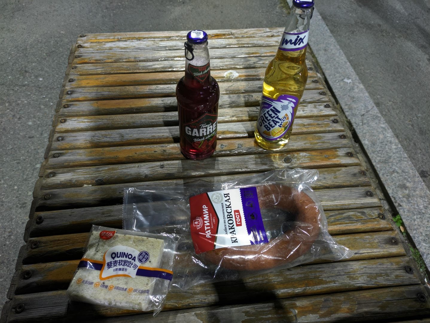 Two bottles of drinks and two kinds of food placed on a wooden table. One bottle is a red 'Garrafa Black' drink, and the other is a yellow 'Green Bean Mix' drink. The food packaging on the left reads 'QUINOA', and the food on the right is a round sausage.