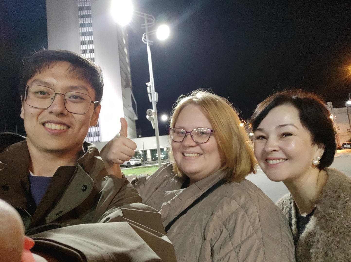 Three people posing for a photo outdoors at night. The person on the left wears glasses, a brown coat, and gives a thumbs-up. The person in the middle wears clear glasses, a light-colored coat, and smiles. The person on the right has shorter hair, wears a dark coat, and also smiles. Buildings and streetlights are visible in the background.