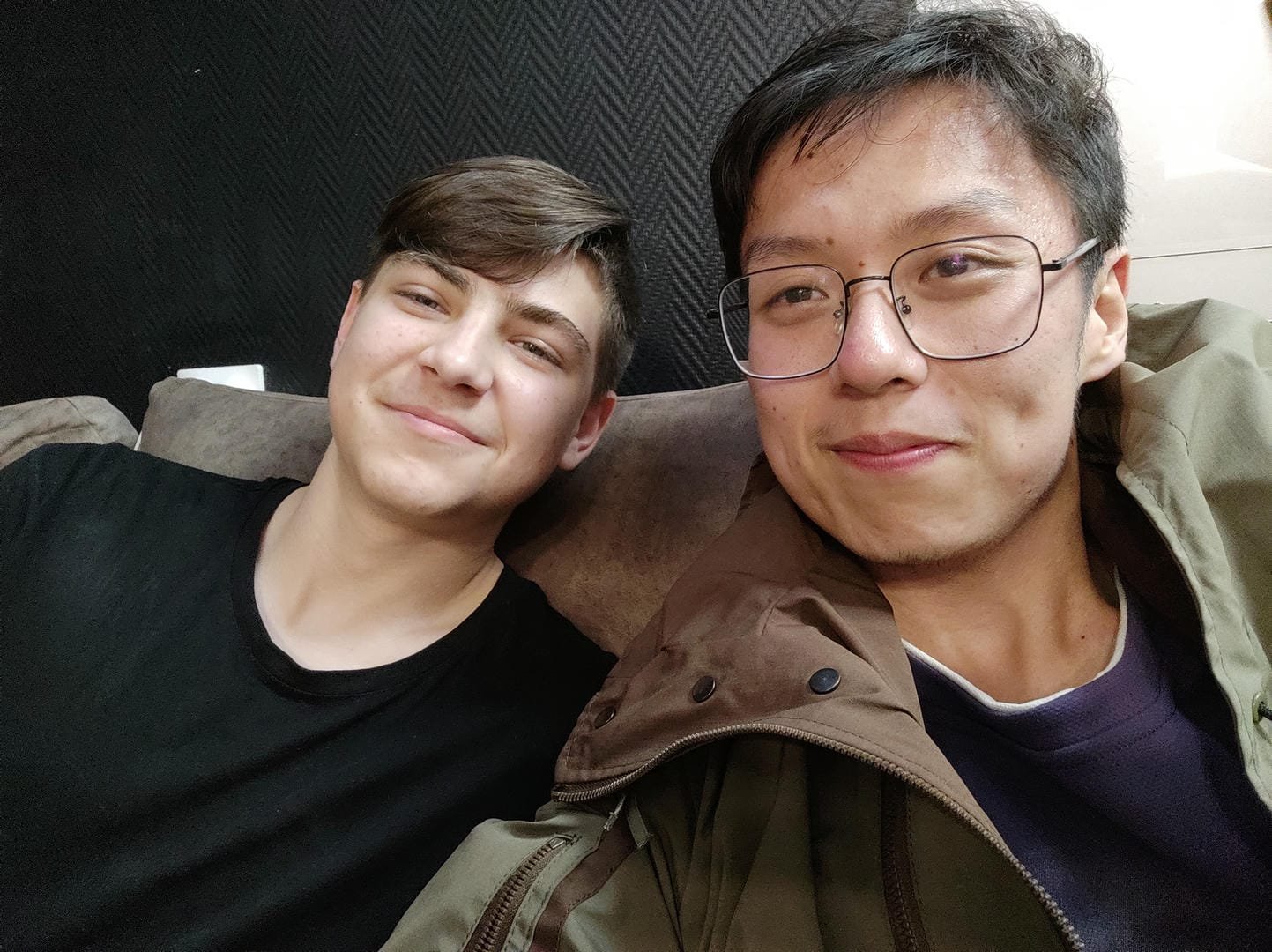 Two people sitting on a sofa; the person on the left is wearing a black top, and the person on the right is wearing glasses and a brown coat.