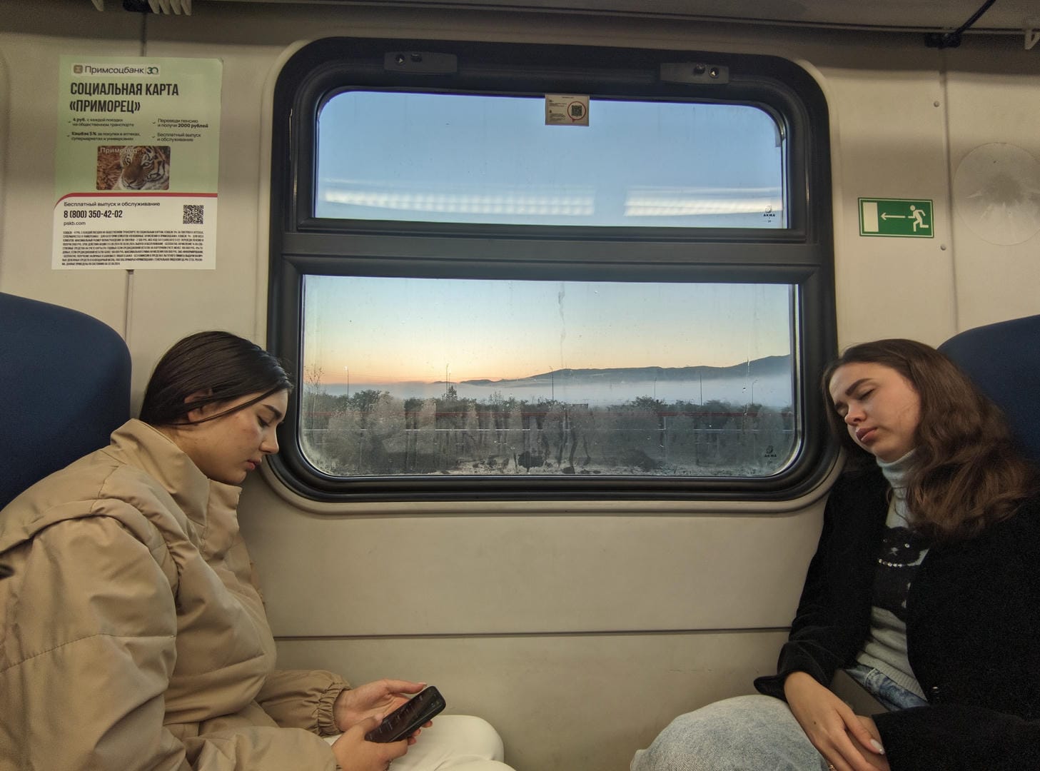 Two women sitting by a train window; the woman on the left is playing with her phone, while the woman on the right is leaning back against the seat to rest.