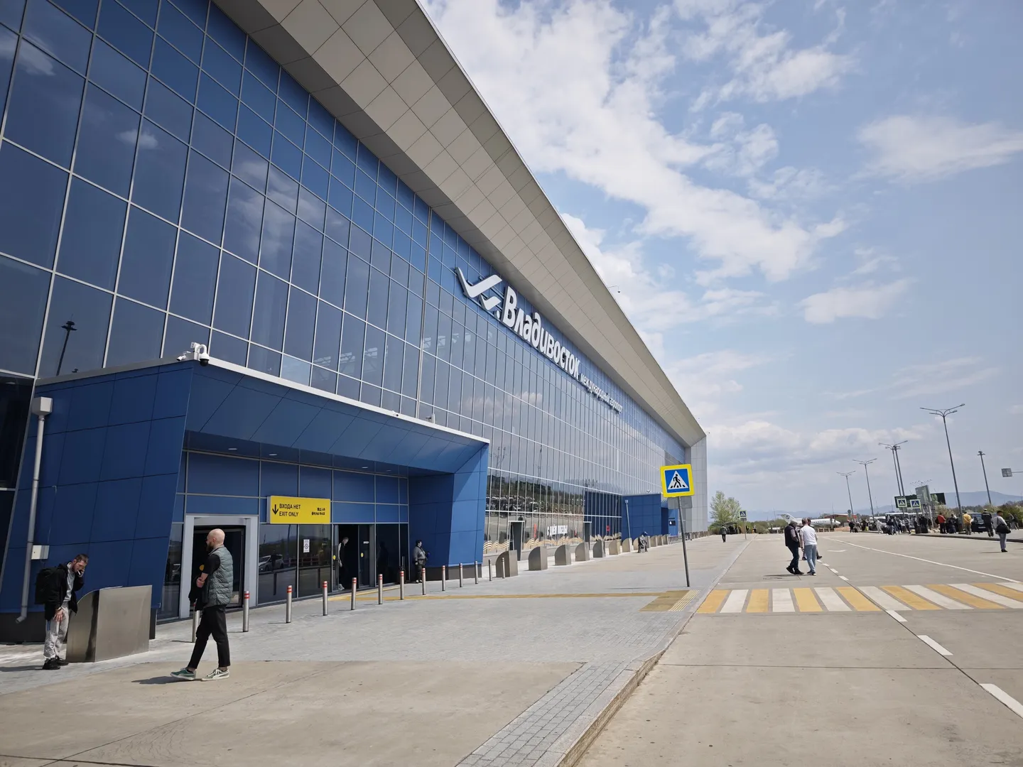 The exterior of a modern building dominated by a blue glass curtain wall. A few people are walking near the entrance under a clear sky on a spacious ground.