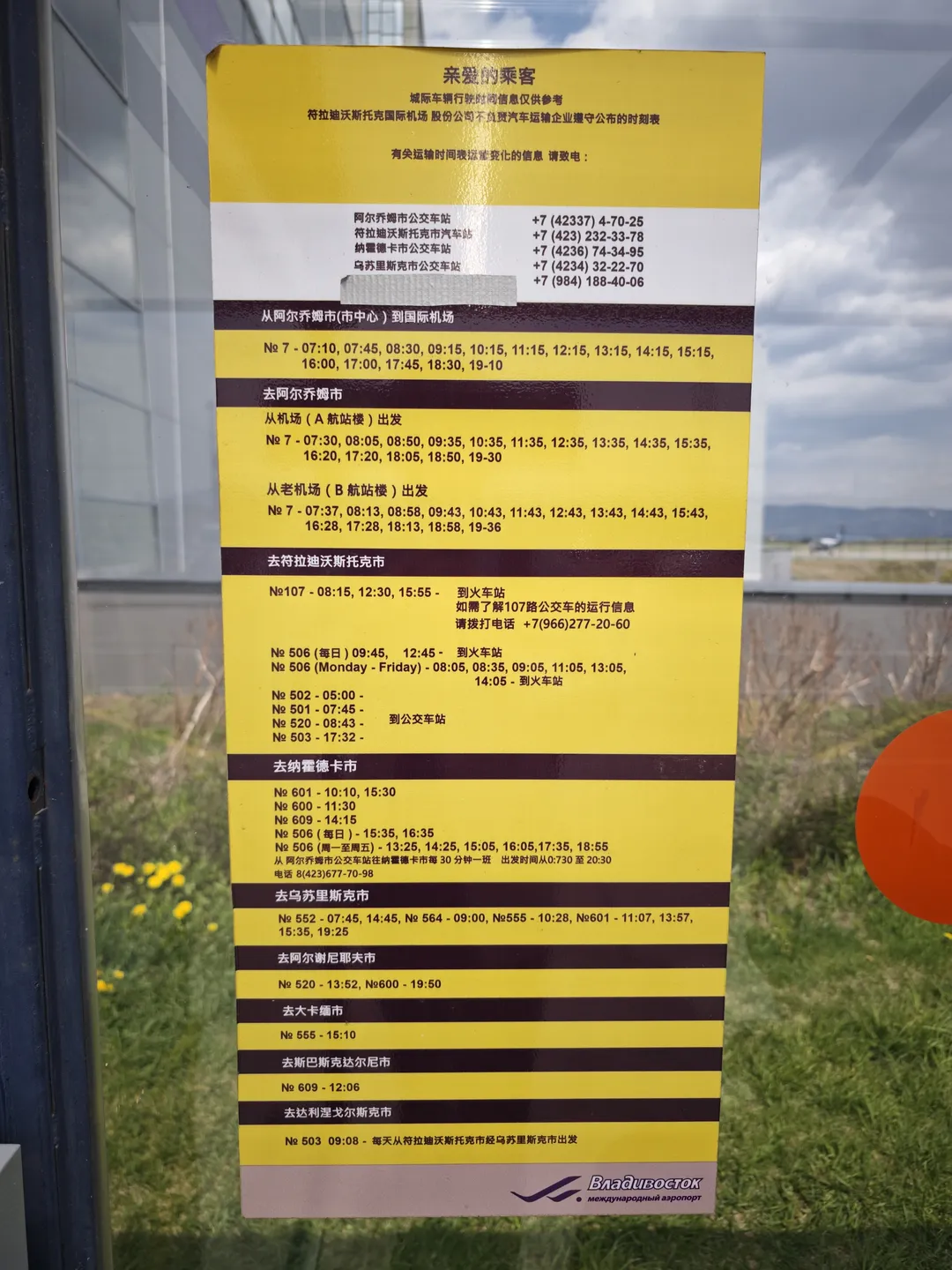 A bus timetable with a yellow background posted on a transparent glass pane. The table details departure times for different routes, against a background of outdoor lawn and distant mountains.