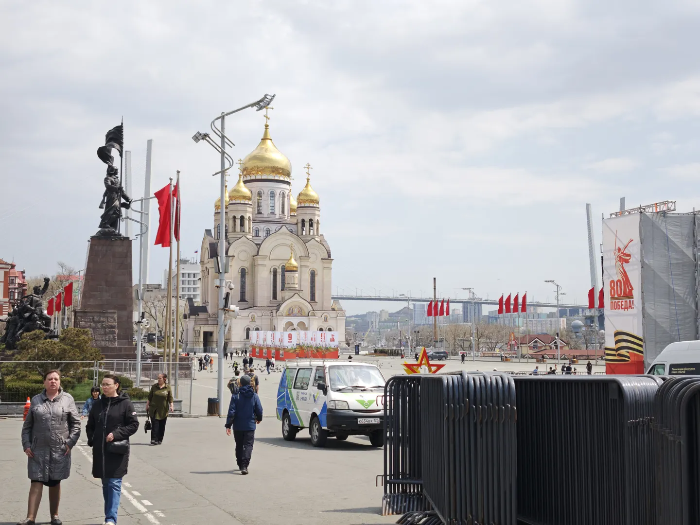 A church with golden domes situated in the center of a square, fronted by red flags. There is a statue to the left of the church and promotional billboards to the right, with pedestrians and vehicles on the square.