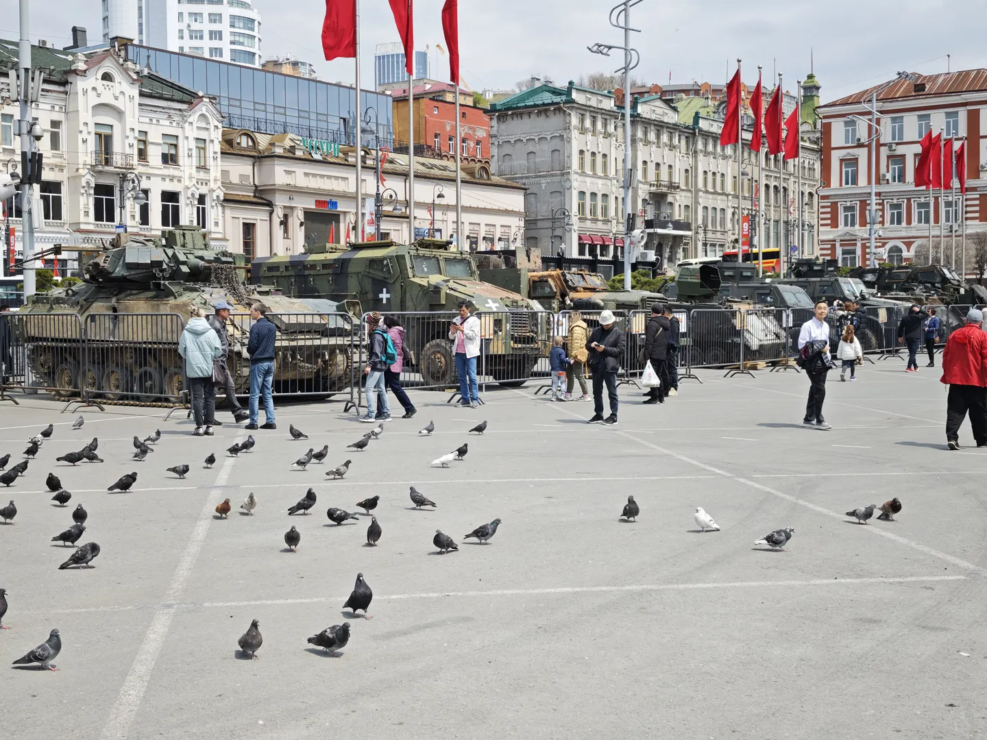 Several military armored vehicles parked on a square, surrounded by fences. Numerous pigeons are on the ground, and people are walking beside the vehicles against a background of multi-story buildings and red flags.