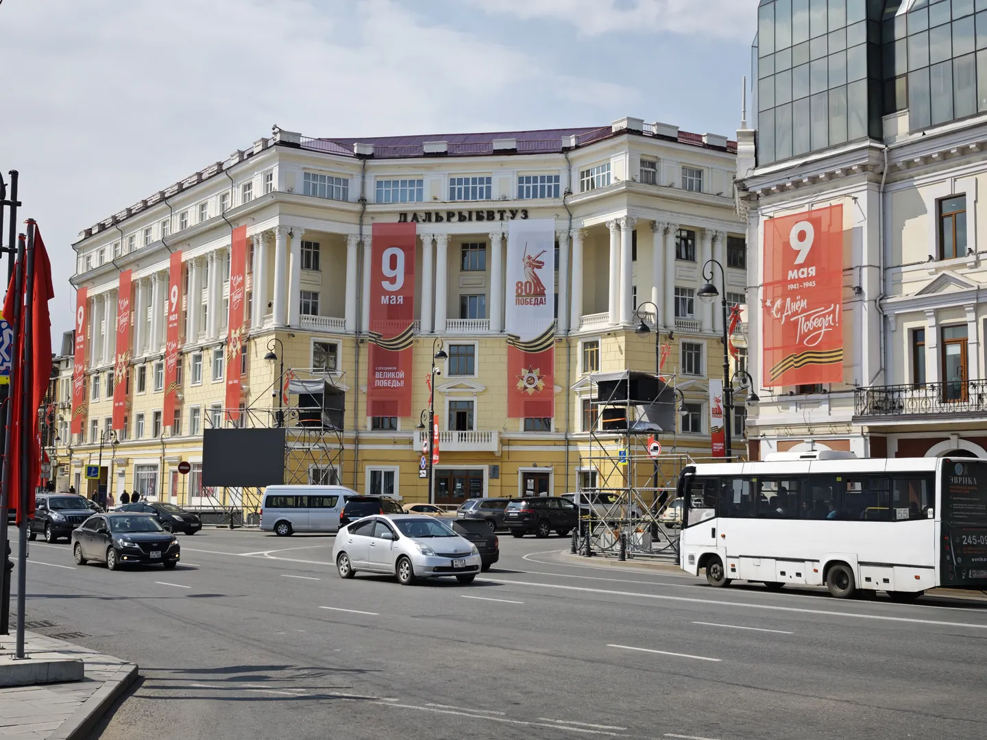A street lined with classical-style buildings decorated with red banners that read '9 мая', with cars and buses driving on the road and neatly arranged windows on the facades.