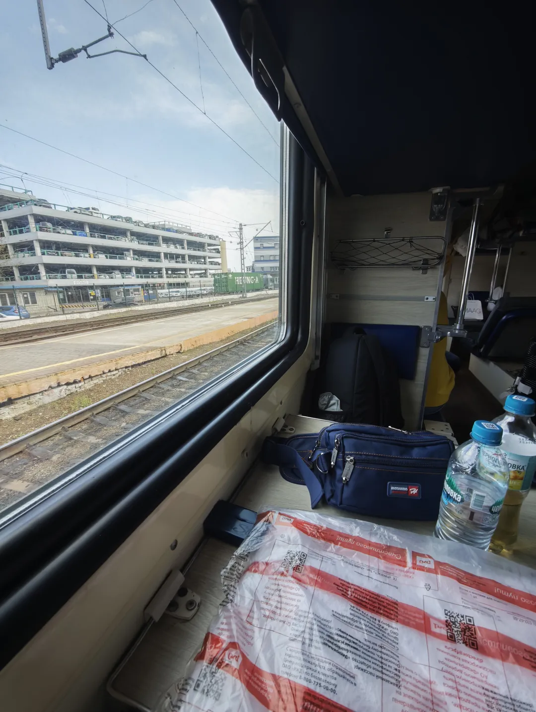 Interior of a train carriage, where railway tracks and a multi-story parking lot are visible outside the window. A blue bag and a water bottle are placed by the window, and luggage racks and seats are inside.