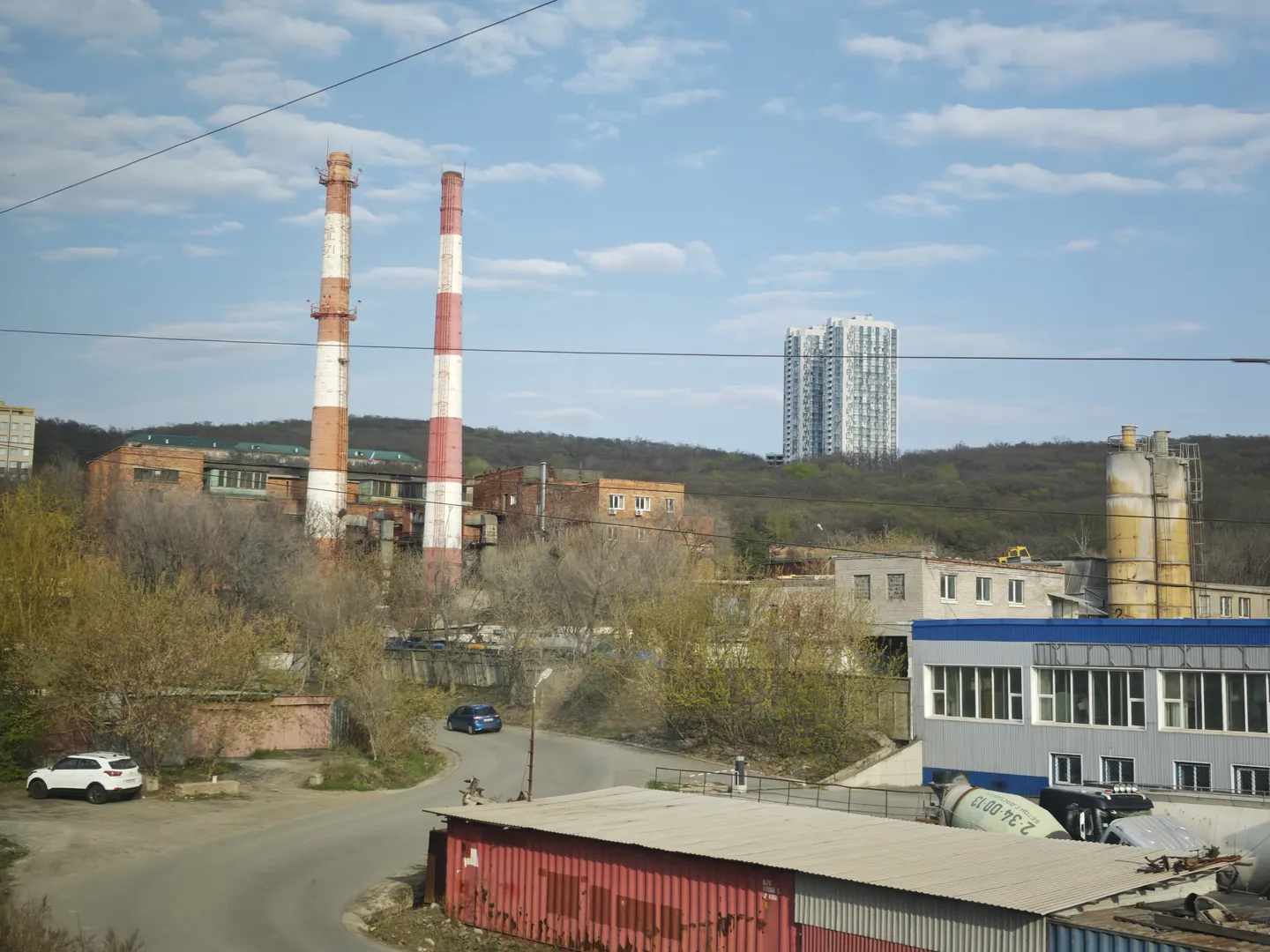 An industrial area scene featuring two tall red-and-white striped chimneys, with a road and parked vehicles in the foreground, and trees alongside high-rise residential buildings in the background.