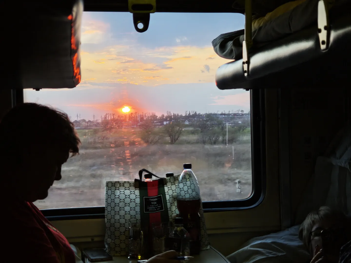 The setting sun is slowly going down outside the window, painting the sky in orange-red. Inside the compartment, a person sits sideways in front of a bottle of drink and a branded shopping bag, while someone lies on the opposite berth with some belongings nearby.
