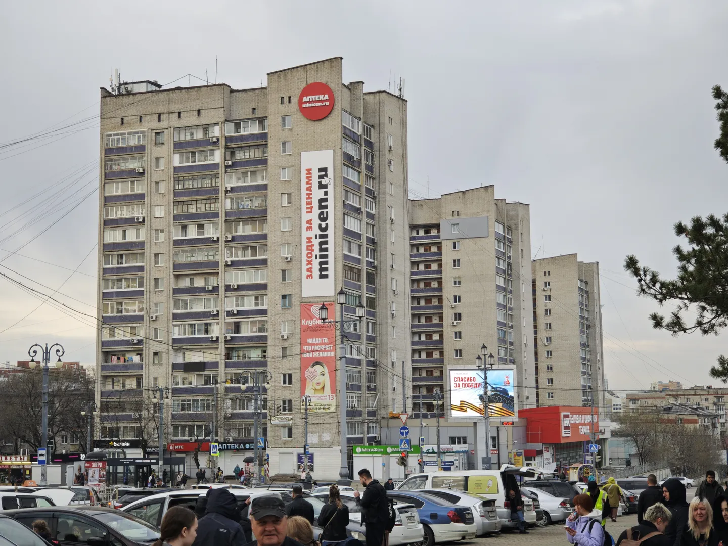 An old high-rise building with a grey facade and neatly arranged windows. Multiple billboards are mounted on the building, with a street full of parked vehicles in the foreground and many people walking by.