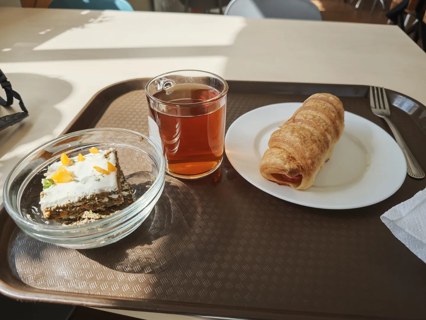A cup of tea, a croissant, and a slice of cake served on a tray. The tea cup is transparent, the croissant is golden brown, and the cake is decorated with cream and orange slices. The brown tray is placed on a white table.